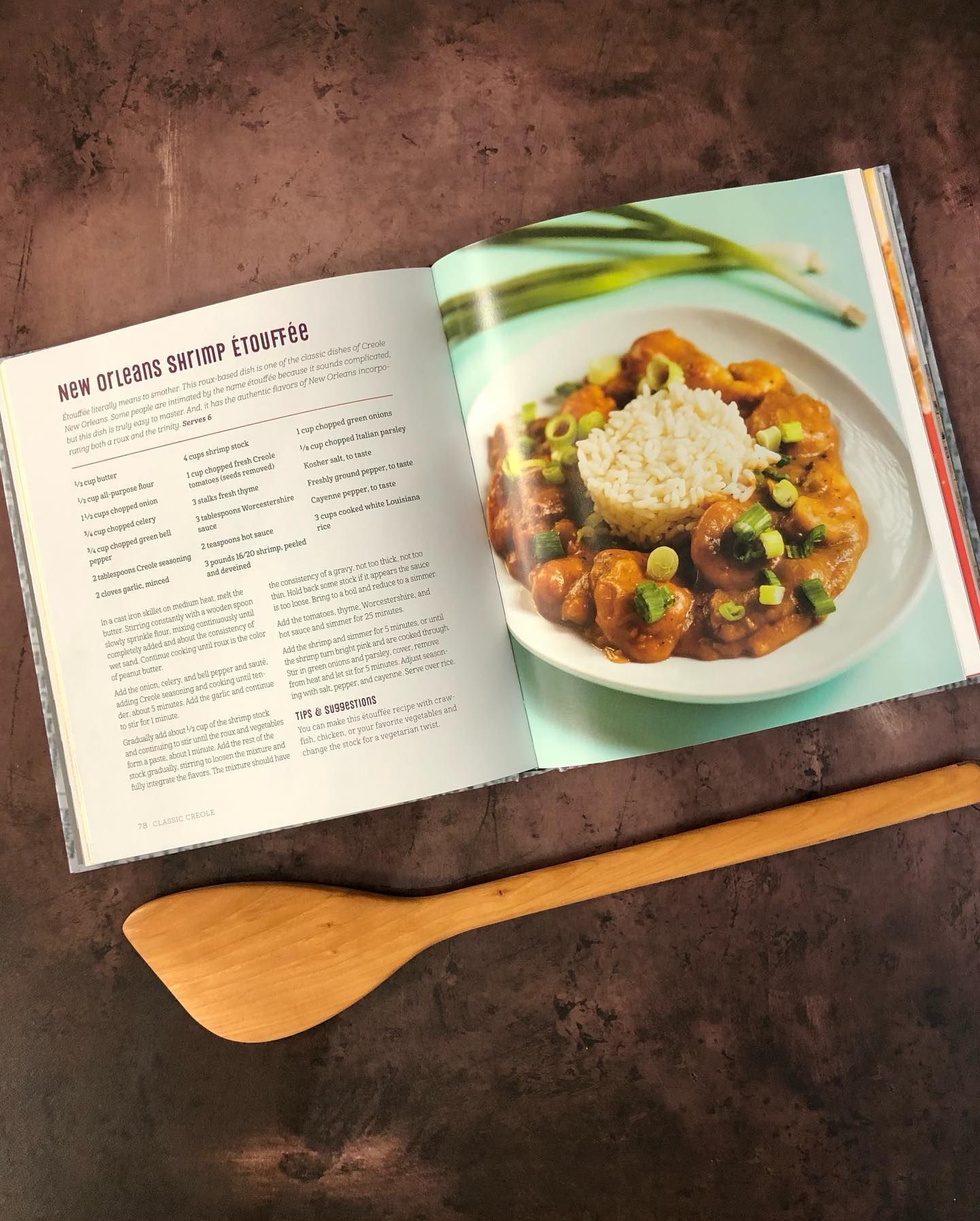 An open cookbook page featuring a recipe for shrimp curry with rice, with a wooden spatula lying on a dark surface.