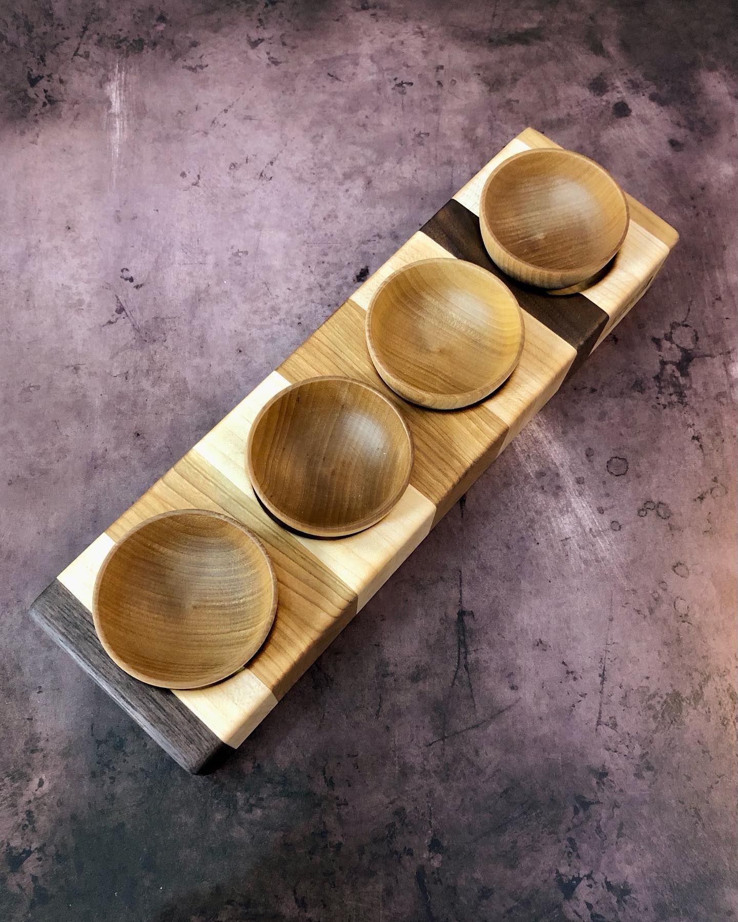 A wooden server with four circular bowls carved into alternating stripes of light and dark wood on a mottled purple background.