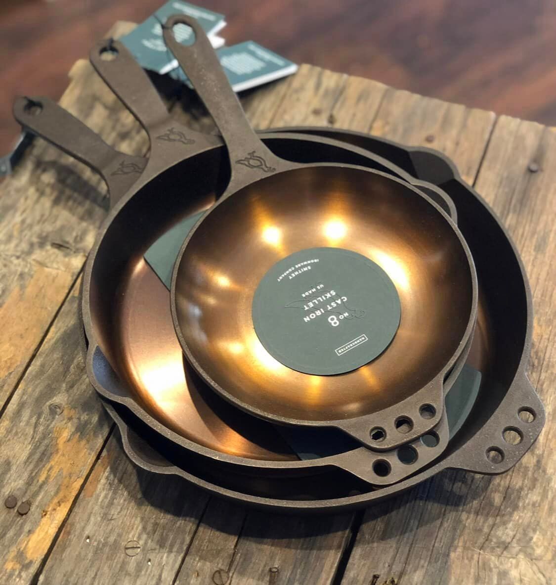 Three nested, bronze-colored cast iron skillets with handles rest on a rustic wooden table.