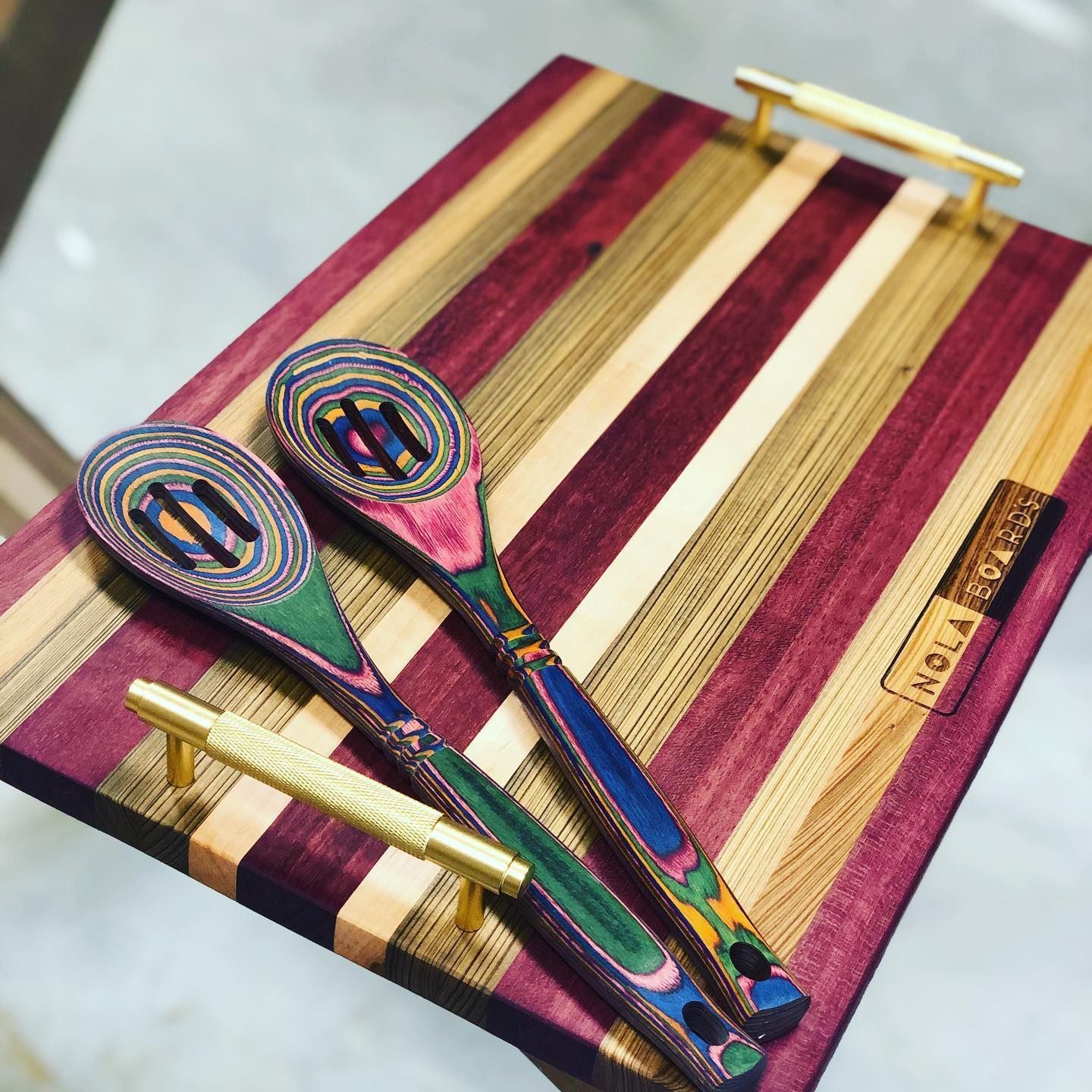 A striped, multicolored wooden serving tray with gold handles and two matching slotted spoons resting on top.
