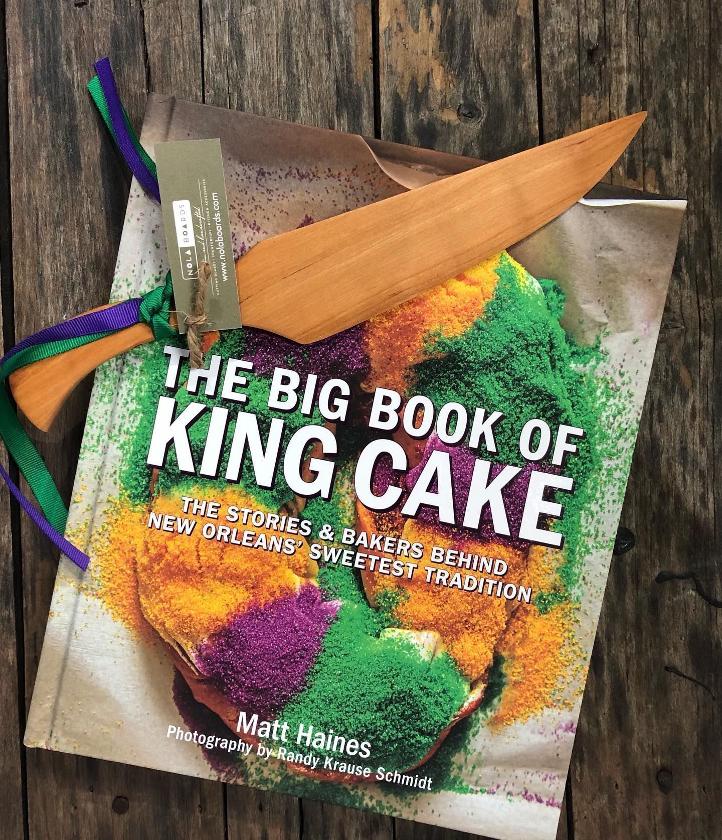 A book titled The Big Book of King Cake, featuring a colorful sugar-dusted cake, with a wooden knife tied by ribbons.