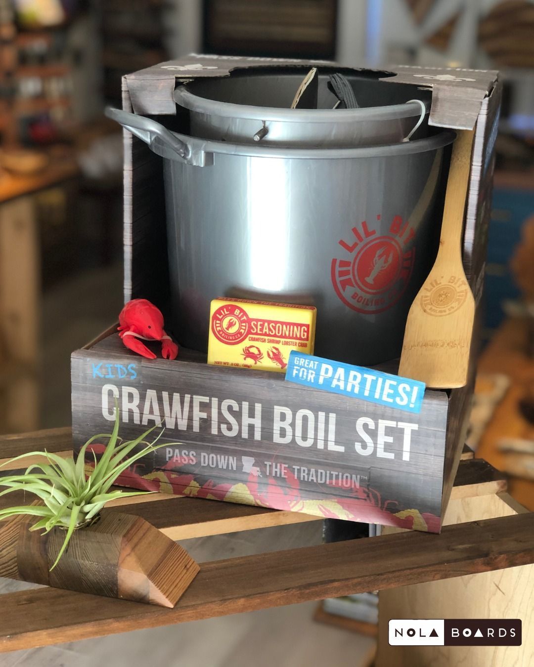 A Nola Boards boxed crawfish boil set including a silver pot, seasoning, a wooden paddle, and a small crawfish toy.
