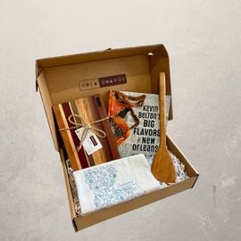 Open cardboard gift box with books, a blue-wrapped item, and a wooden spoon on a light surface