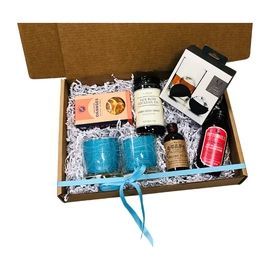 Gift box with assorted spa items, including bottles, jars, and blue candles on shredded paper.