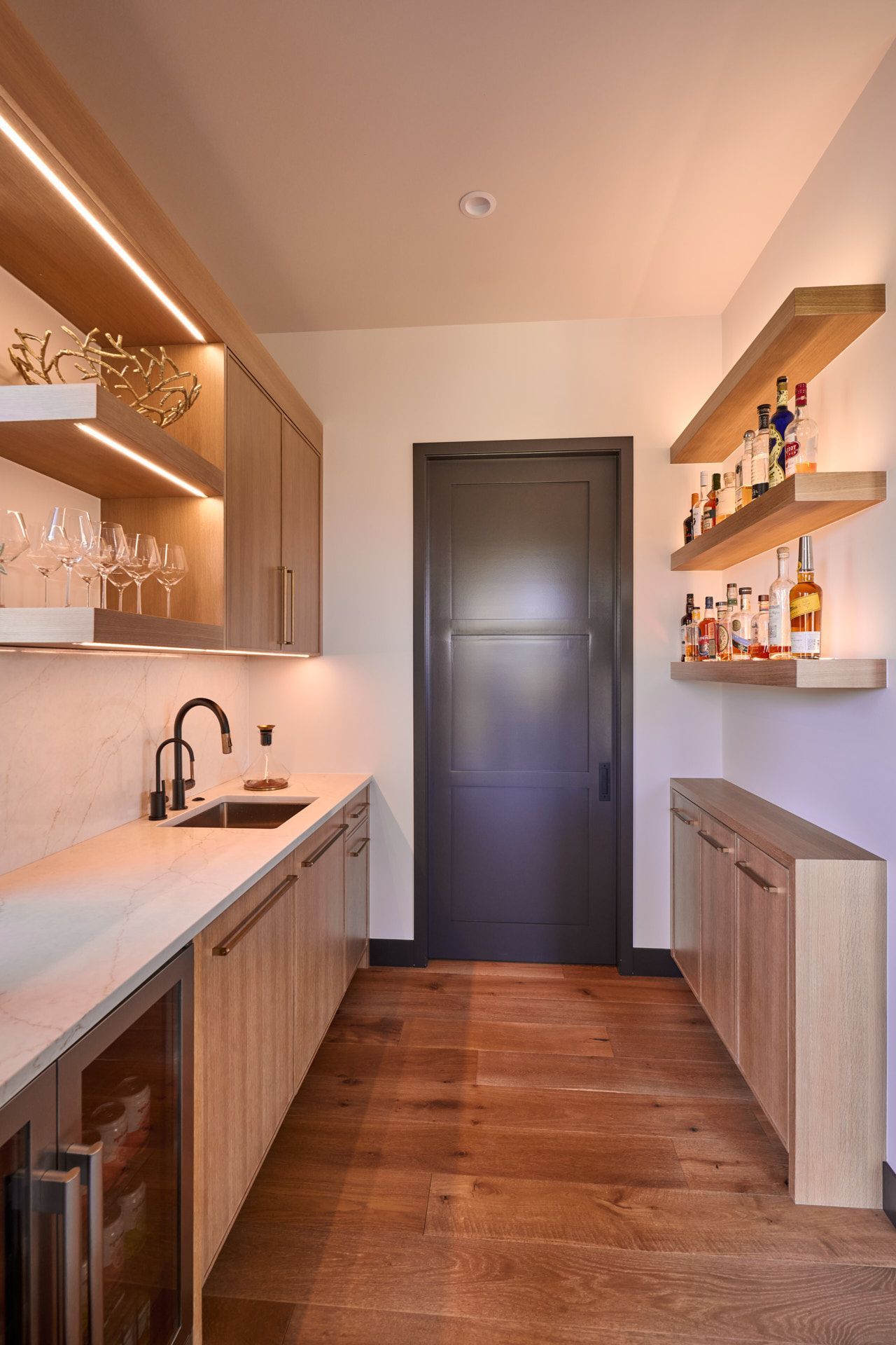 A kitchen with a sink , refrigerator , shelves and bottles on the shelves.