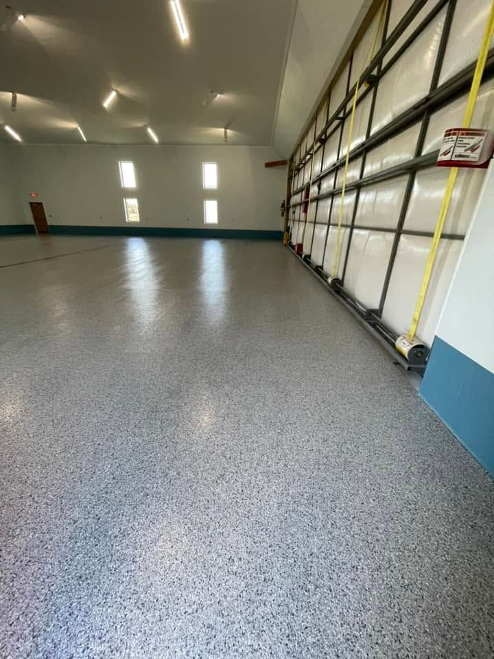 Empty indoor warehouse with speckled gray floor, white walls, ceiling lights, and tall storage racks along one side