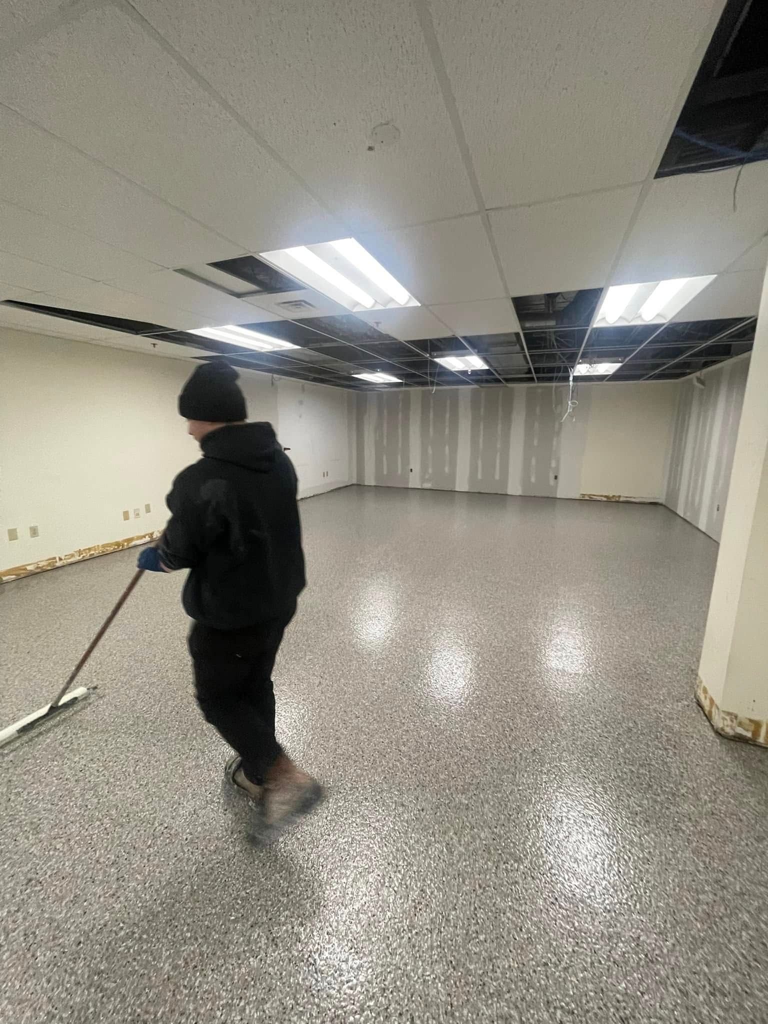 Person in black clothing walking through a large empty indoor room with fluorescent ceiling lights.