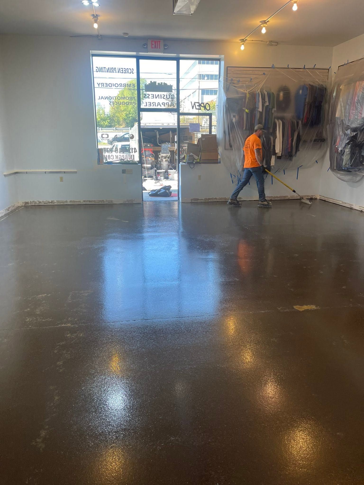 Empty retail showroom with polished concrete floor, bright front windows, and a person in an orange shirt walking inside.