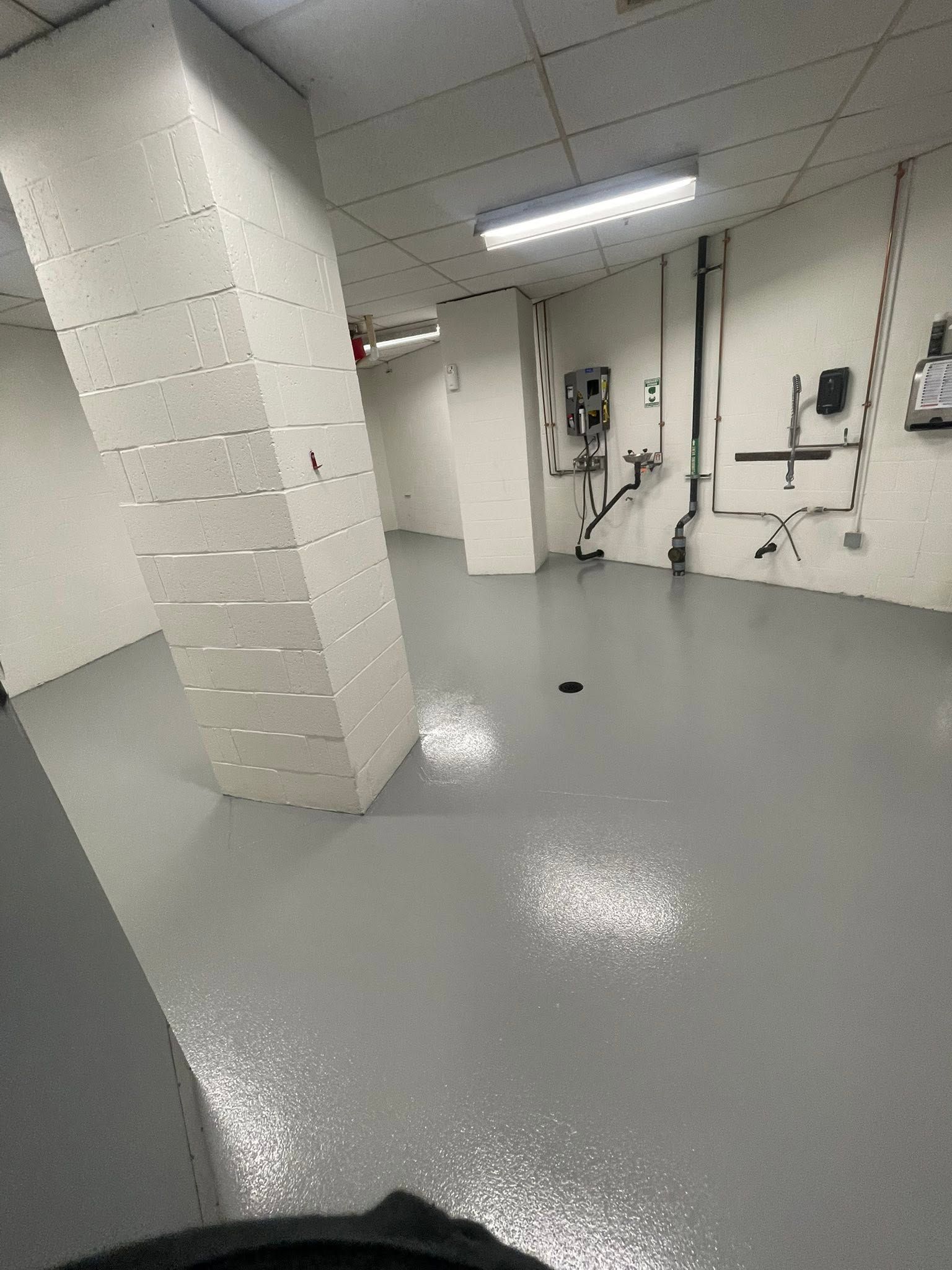 Empty industrial room with gray floor, white walls, ceiling lights, and exposed pipes and equipment.
