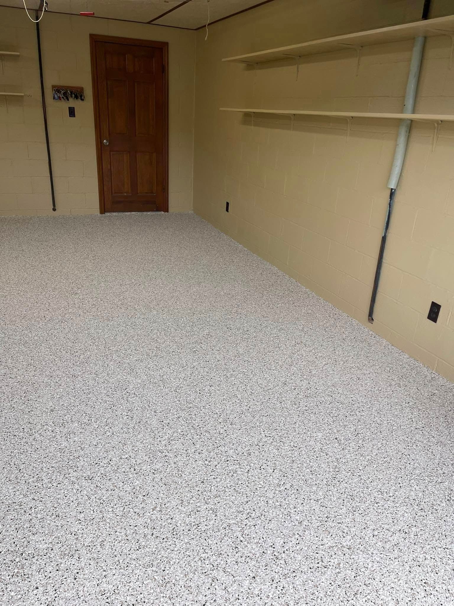 Empty garage with speckled floor, beige walls, wooden door, and wall-mounted shelves.