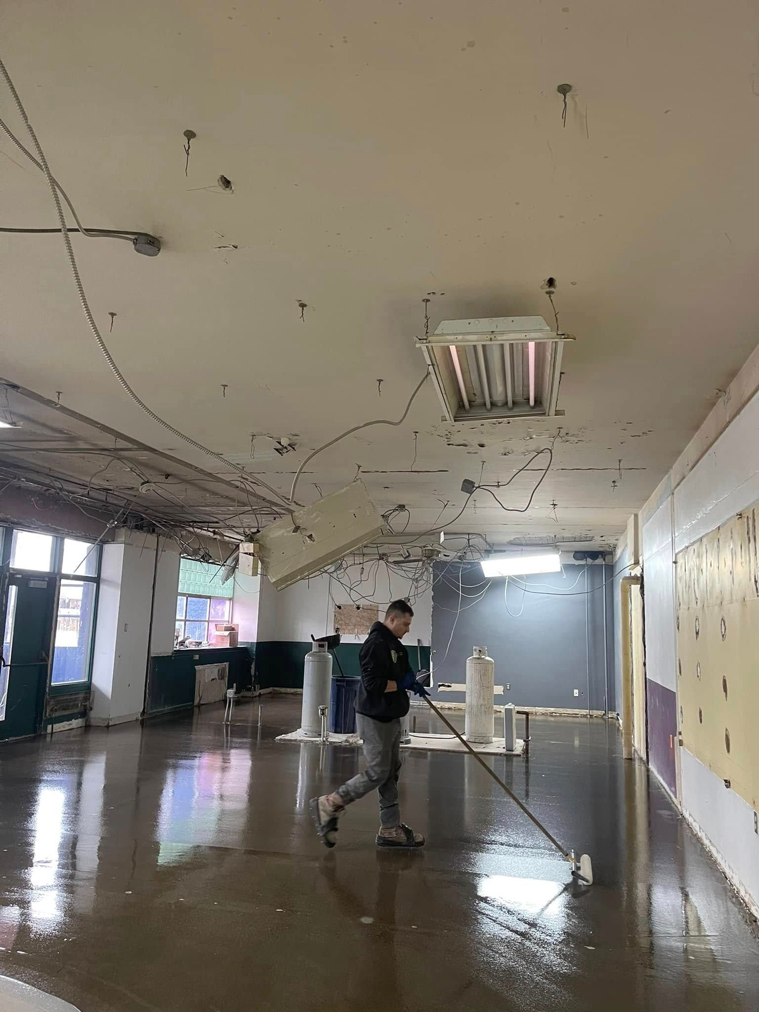 Mop-wet hallway with a worker cleaning near a ceiling light fixture in a damaged building interior.