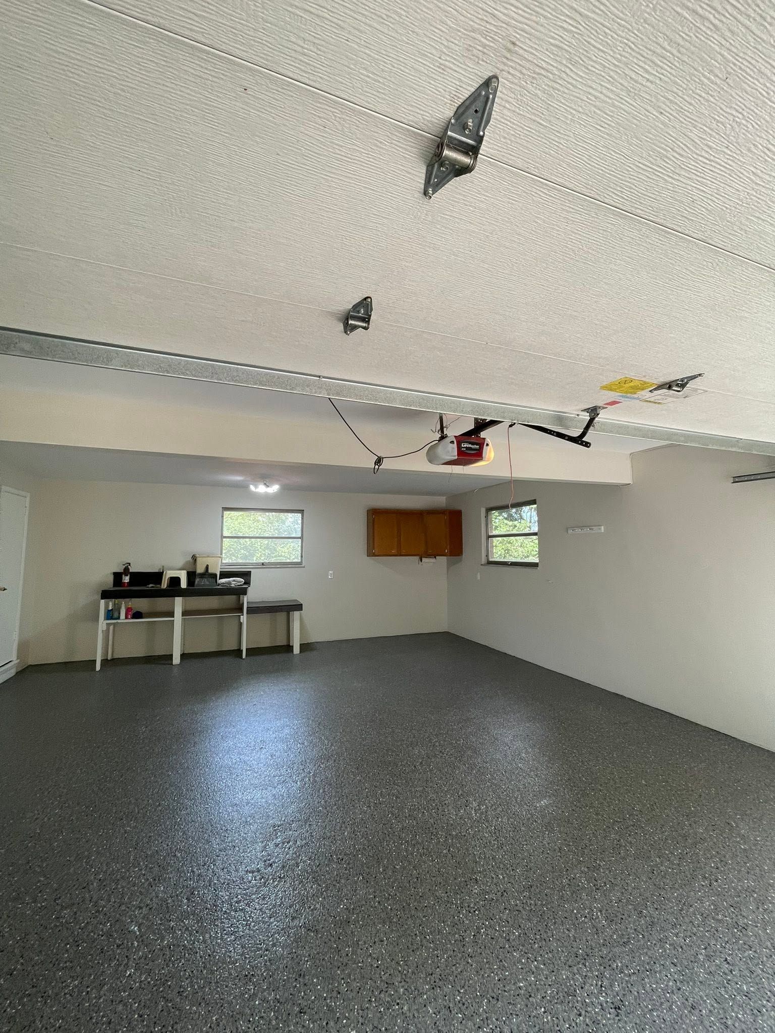Empty garage with gray speckled floor, white walls, overhead lights, and a small worktable by two windows