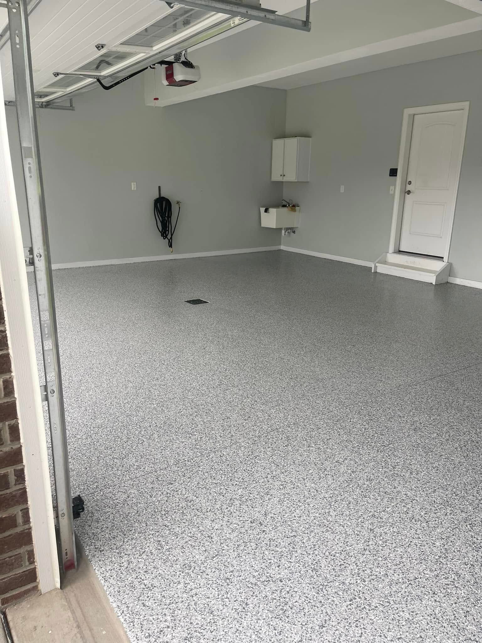 Empty gray garage with speckled epoxy floor, closed white door, and overhead garage door opener.
