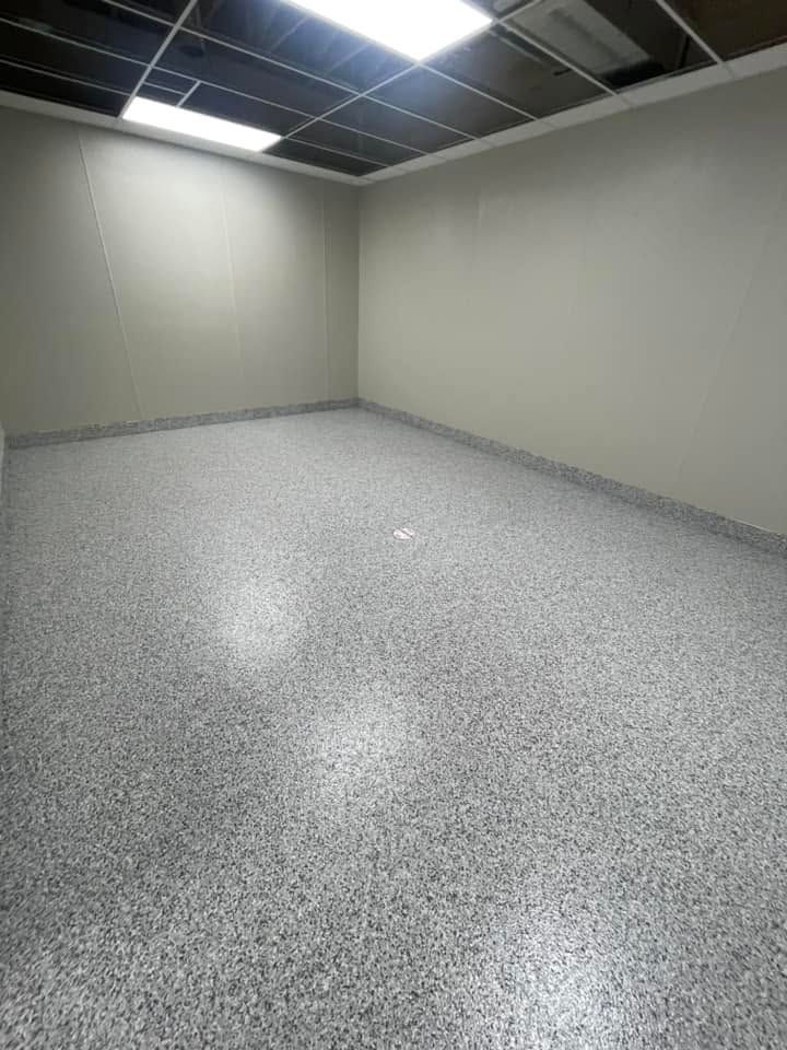 Empty office room with beige walls, speckled gray floor, and a drop ceiling with fluorescent lights