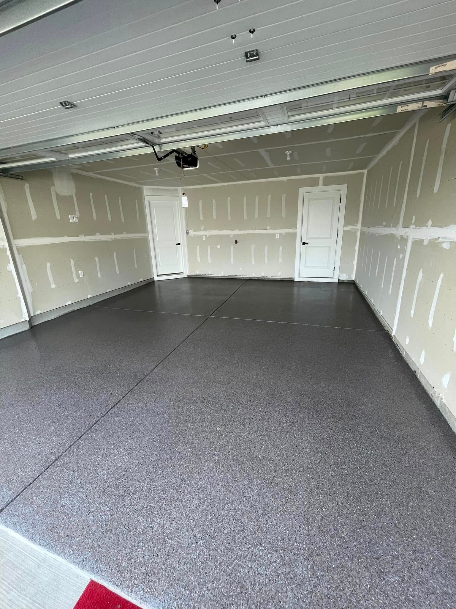 Empty unfinished garage with gray speckled floor, drywall walls, white doors, and overhead lighting.