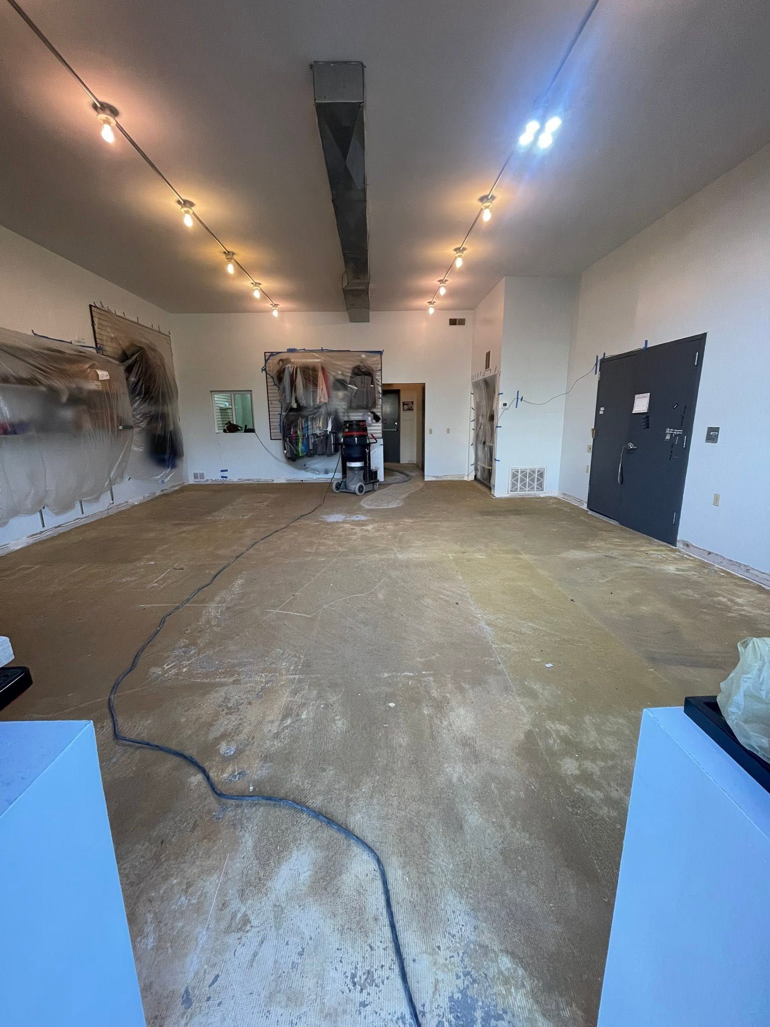 Large empty room under renovation with exposed floor, ceiling lights, and tools near the back wall