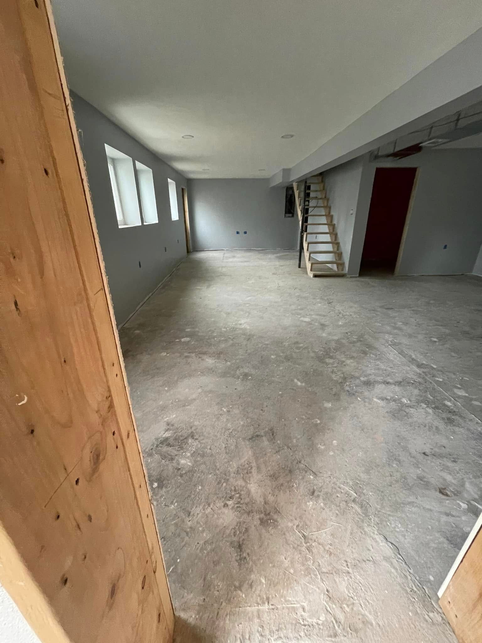Empty unfinished room with gray walls, concrete floor, windows, and a loft ladder near a red door.