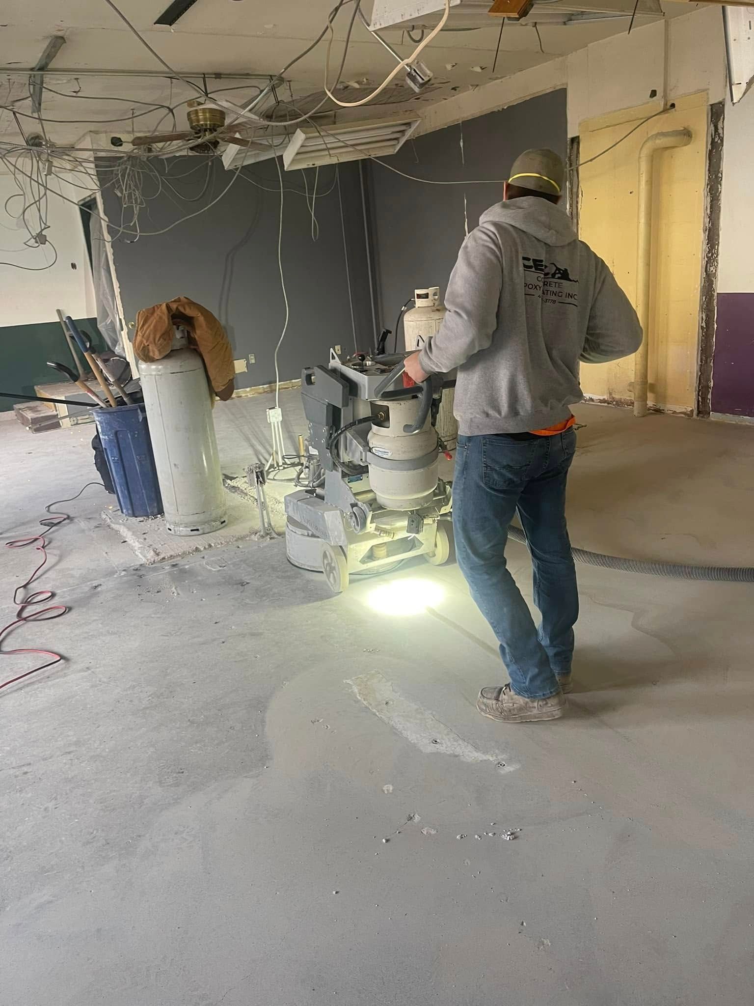 Construction workers using a floor sander in a dusty, unfinished room with exposed ceiling wires
