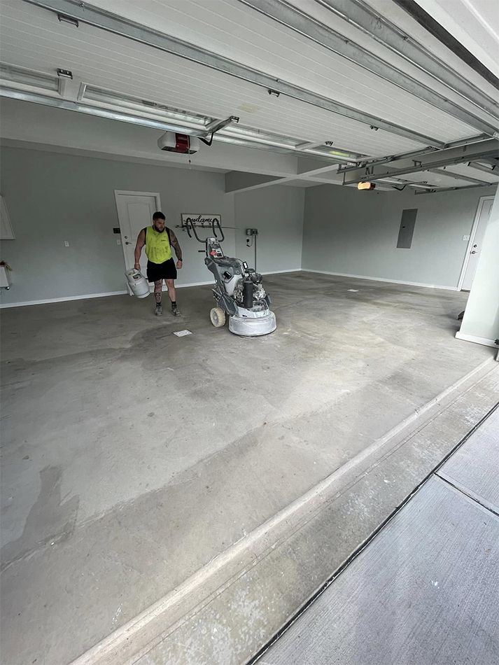 Two people in a garage beside a floor grinding machine, with a concrete floor and open garage door.