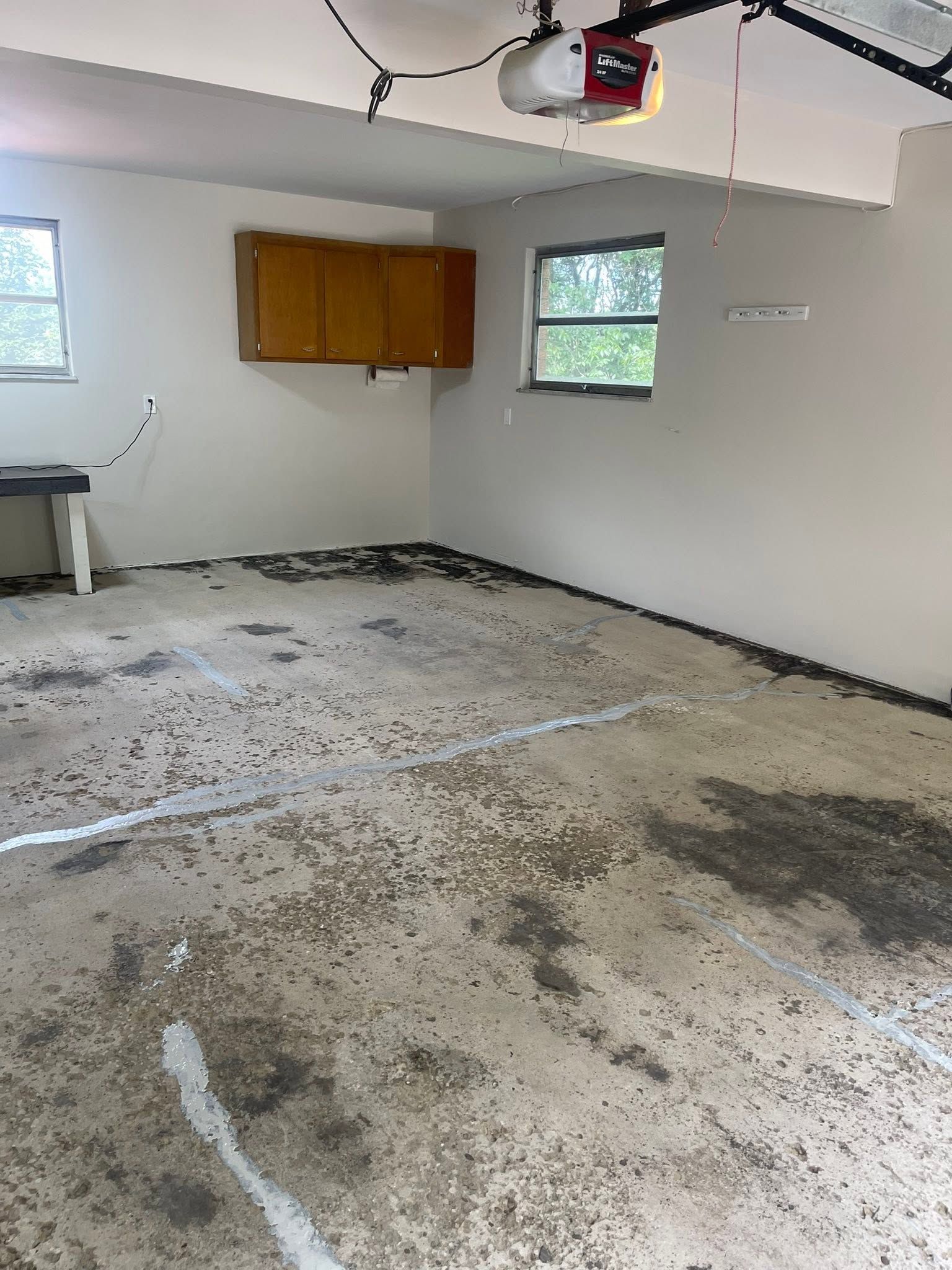 Damaged garage interior with stained concrete floor, white walls, wood cabinets, and a ceiling-mounted opener.