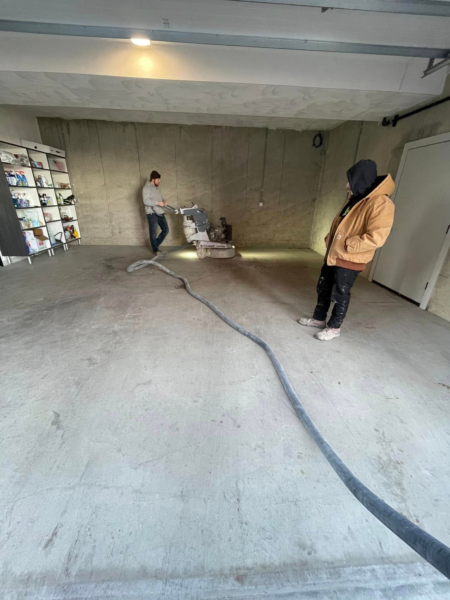 Two workers in a bare concrete room with a hose or cable running across the floor