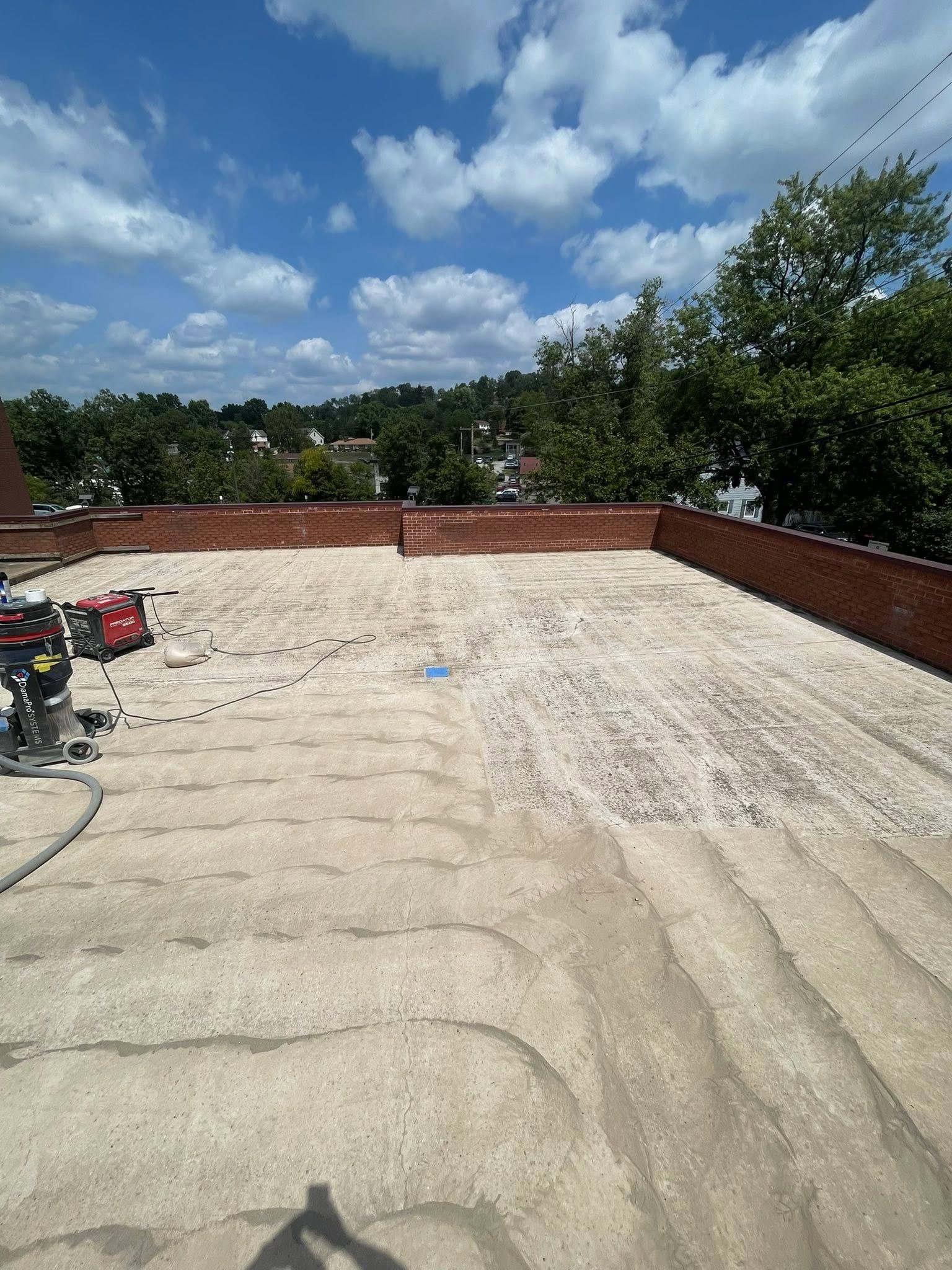 Rooftop with gravel surface, low brick parapet, scattered tools, and trees under a partly cloudy sky