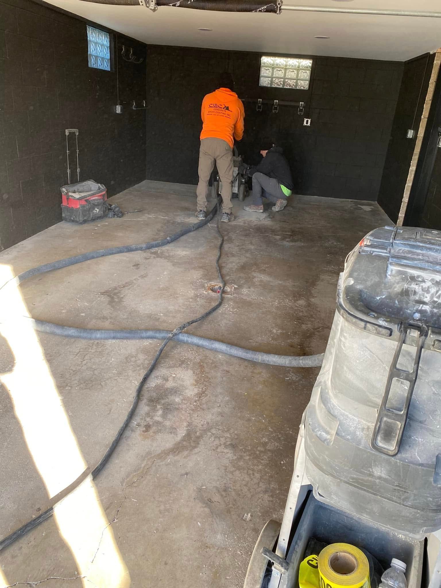 Workers operating a floor grinder in a dark room with hoses and equipment scattered around.