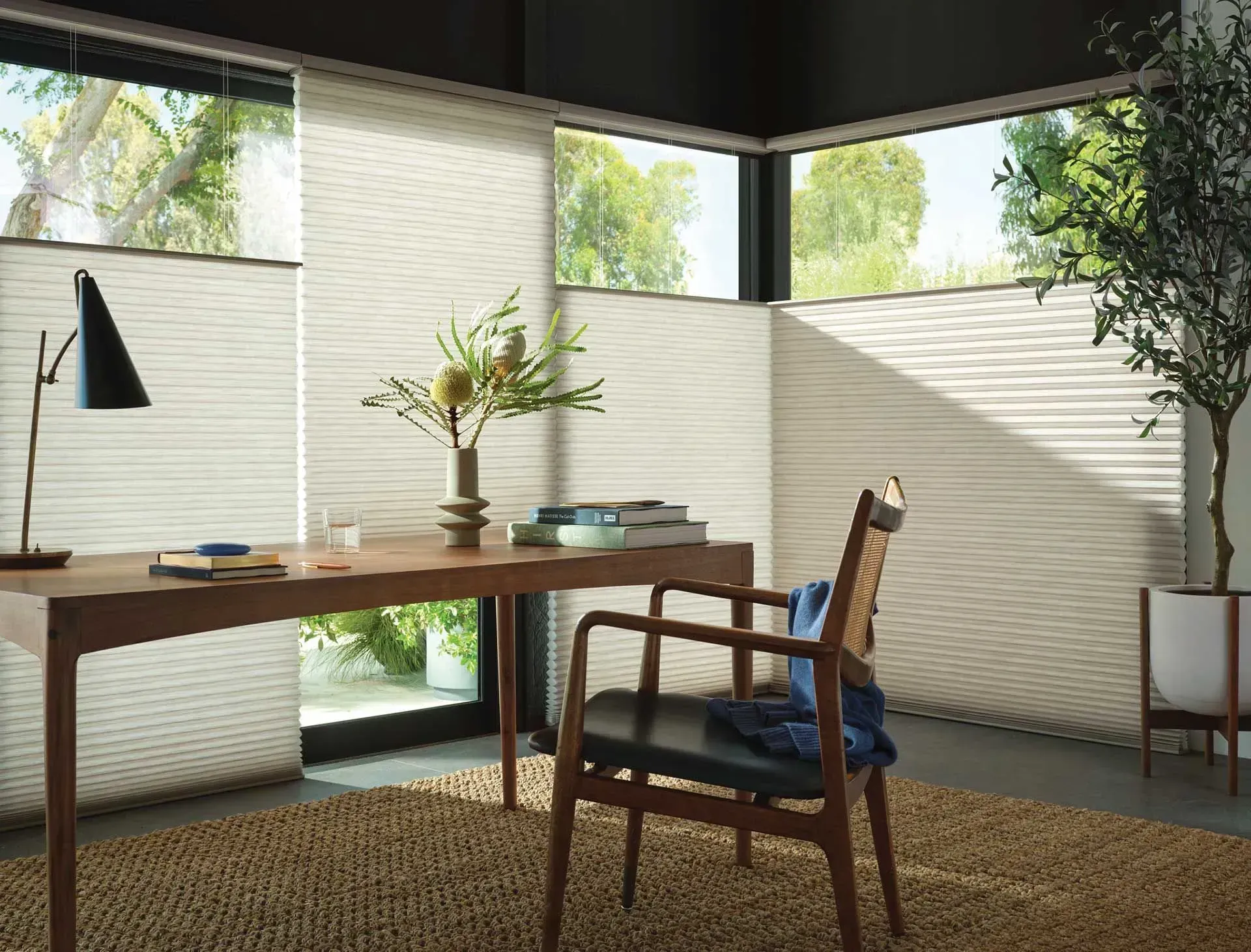 Home office with desk, chair, and beige honeycomb shades; sunlight streams through windows.