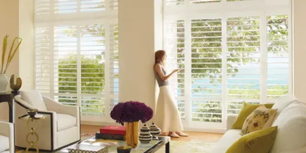 Woman stands near large window, looking outside. White shutters, sea view. Interior with furniture, flowers.