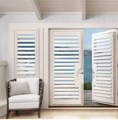 White louvered shutters on a door and window, opened to a view of the ocean. A white chair sits nearby.