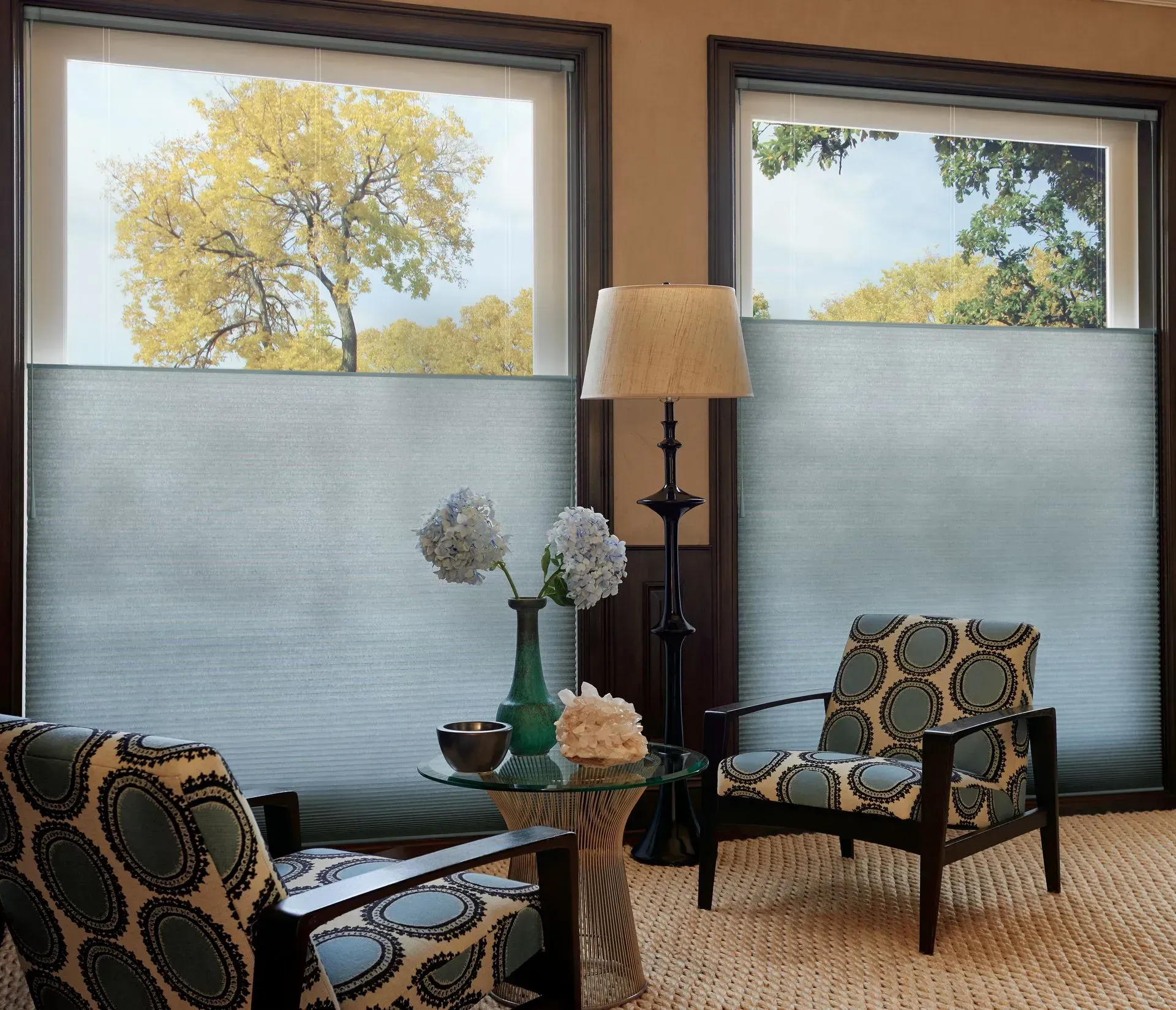 Two windows with top-down, bottom-up cellular shades. Two patterned chairs face each other, with a side table, lamp, and flowers.