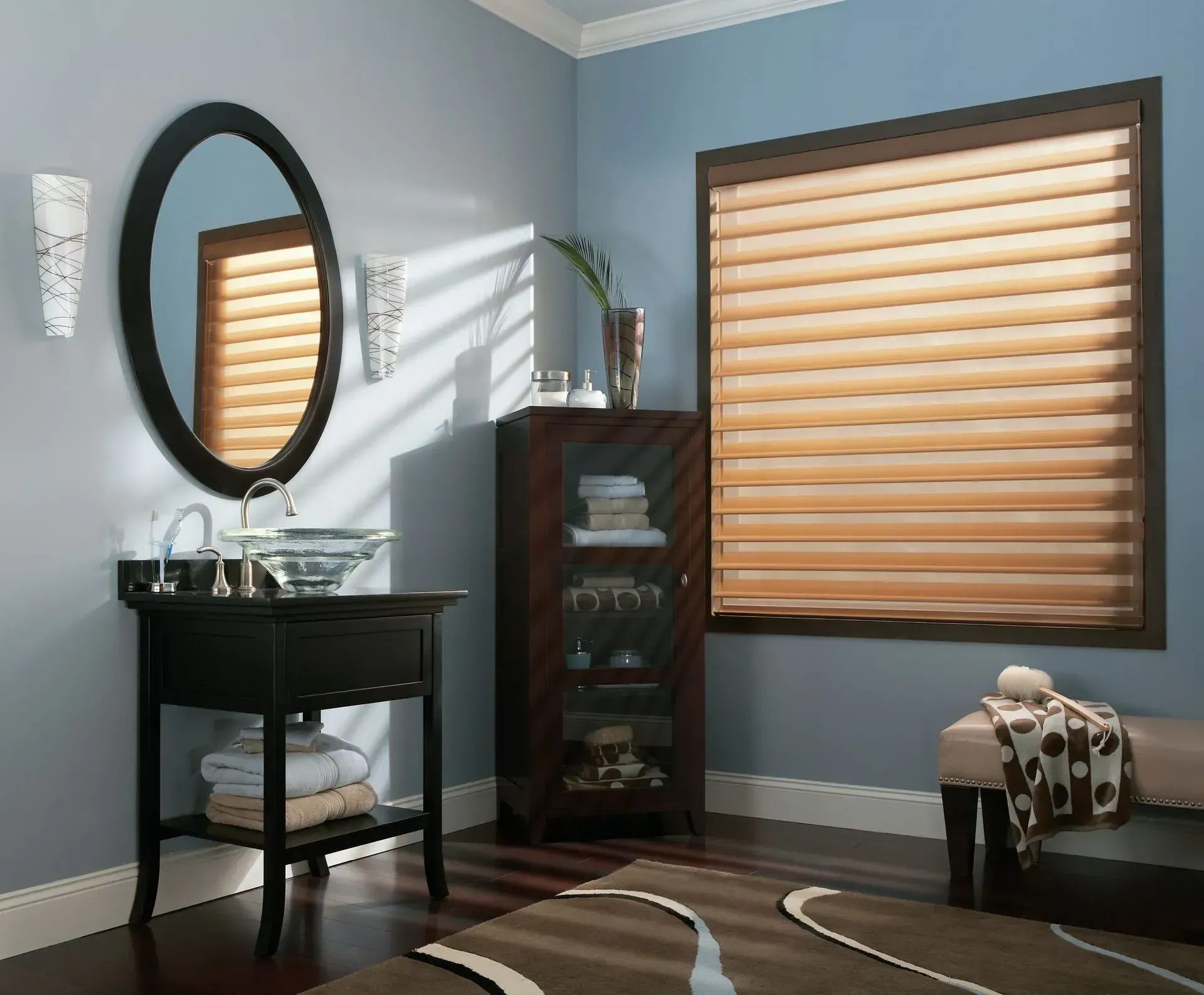 Bathroom with oval mirror, vanity, and window with tan blinds. Blue walls, brown cabinet and rug.