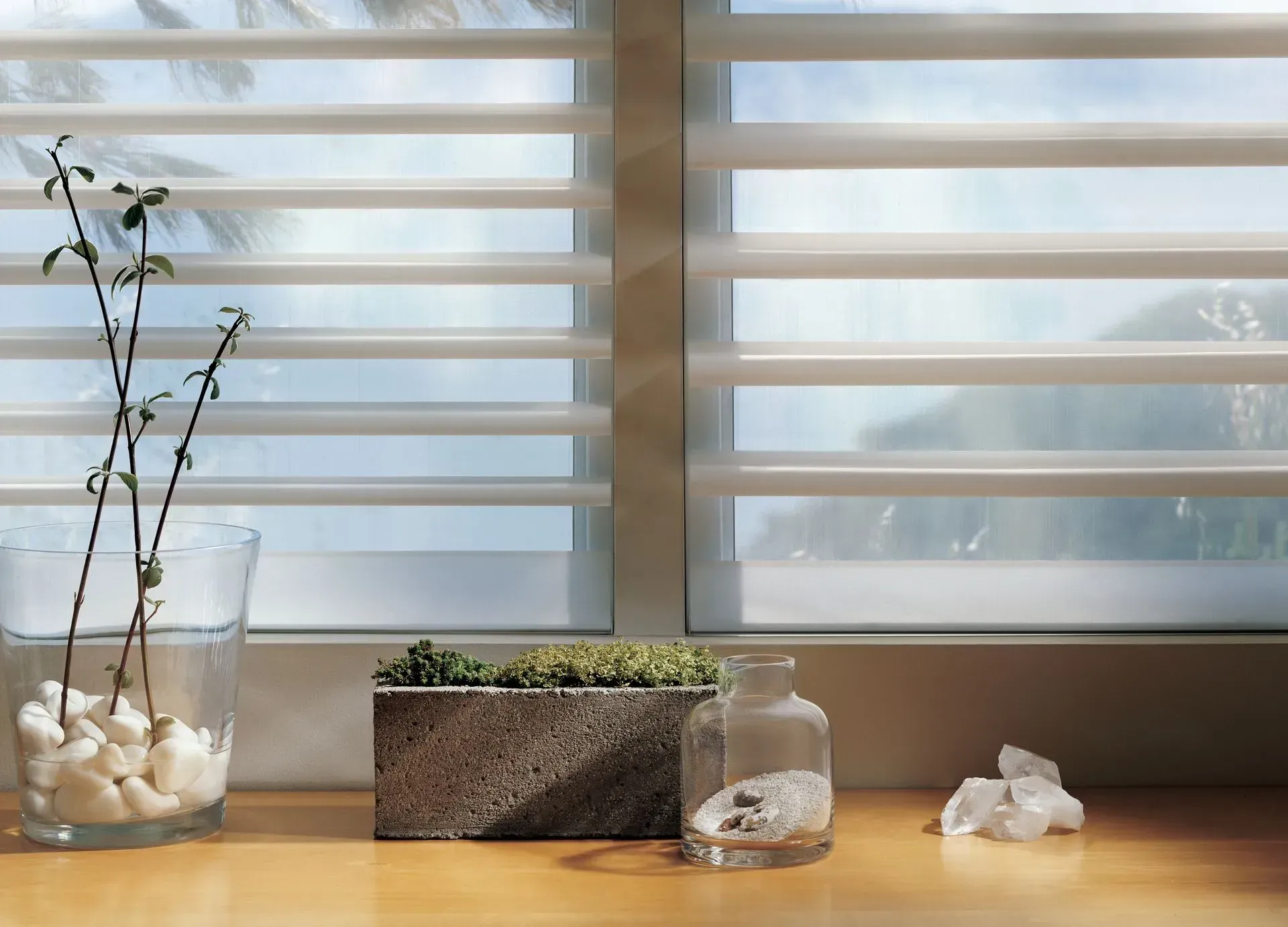 Window with horizontal blinds; still-life display on a wooden sill: vase of stems, concrete planter, glass bottles.