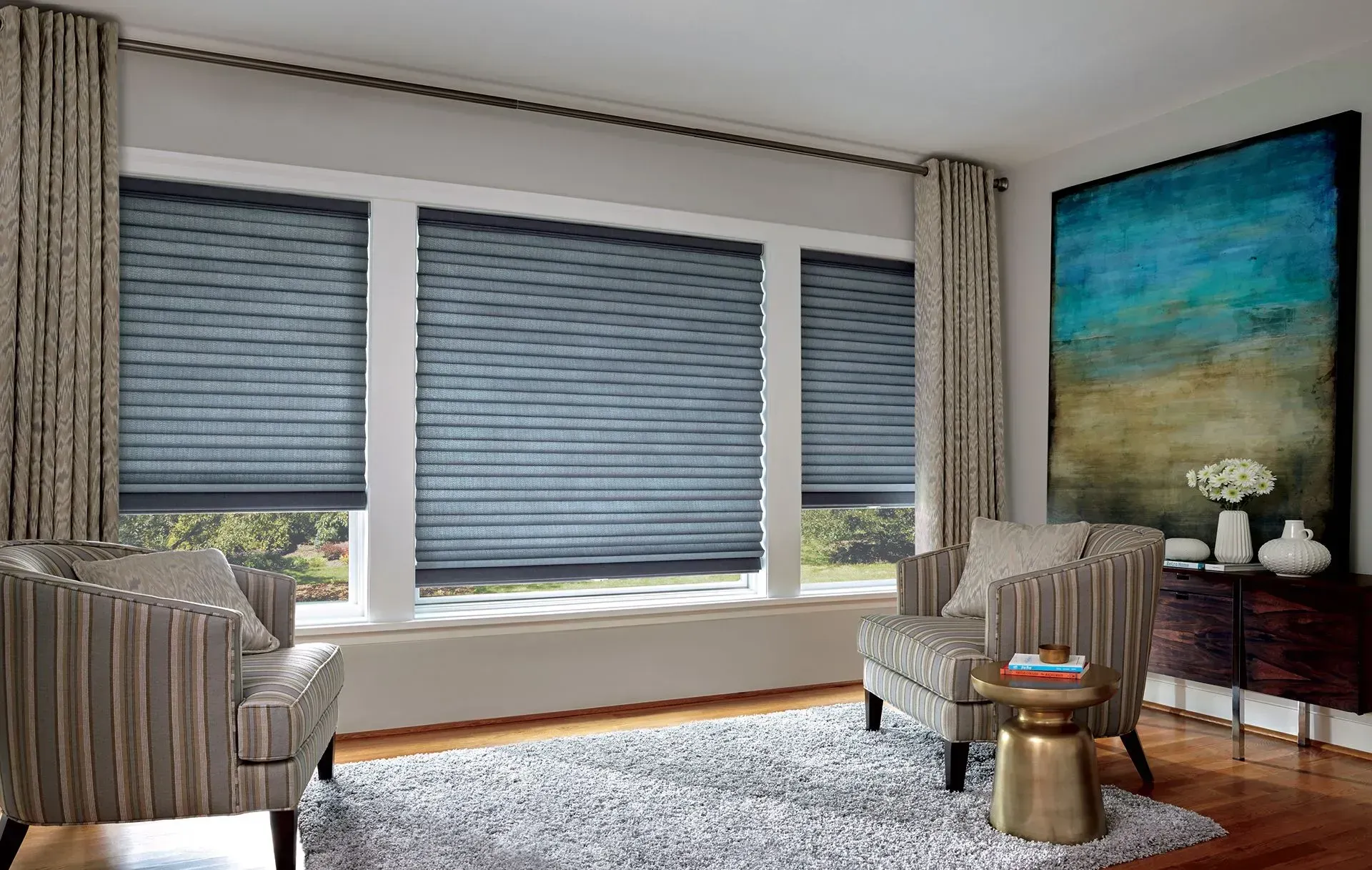 Living room with gray blinds, striped armchairs, large painting, and gold accent table.