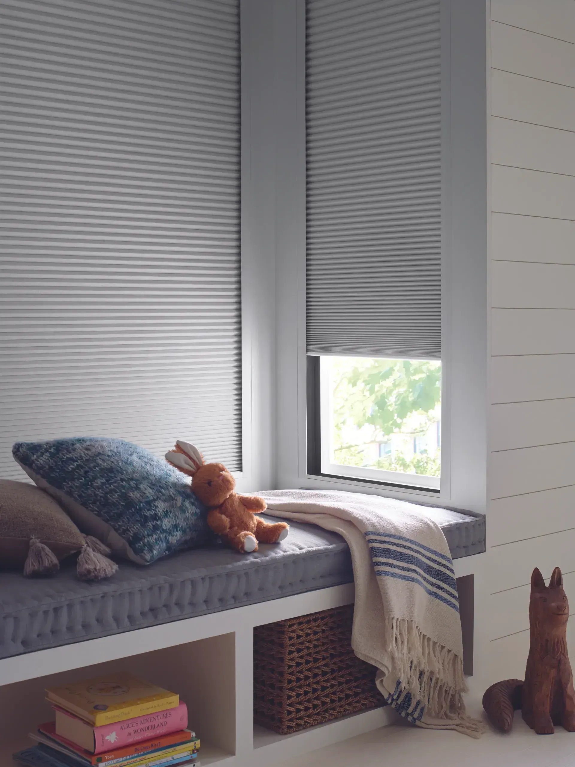 Cozy window seat with gray blinds, pillows, a blanket, and stuffed animals.