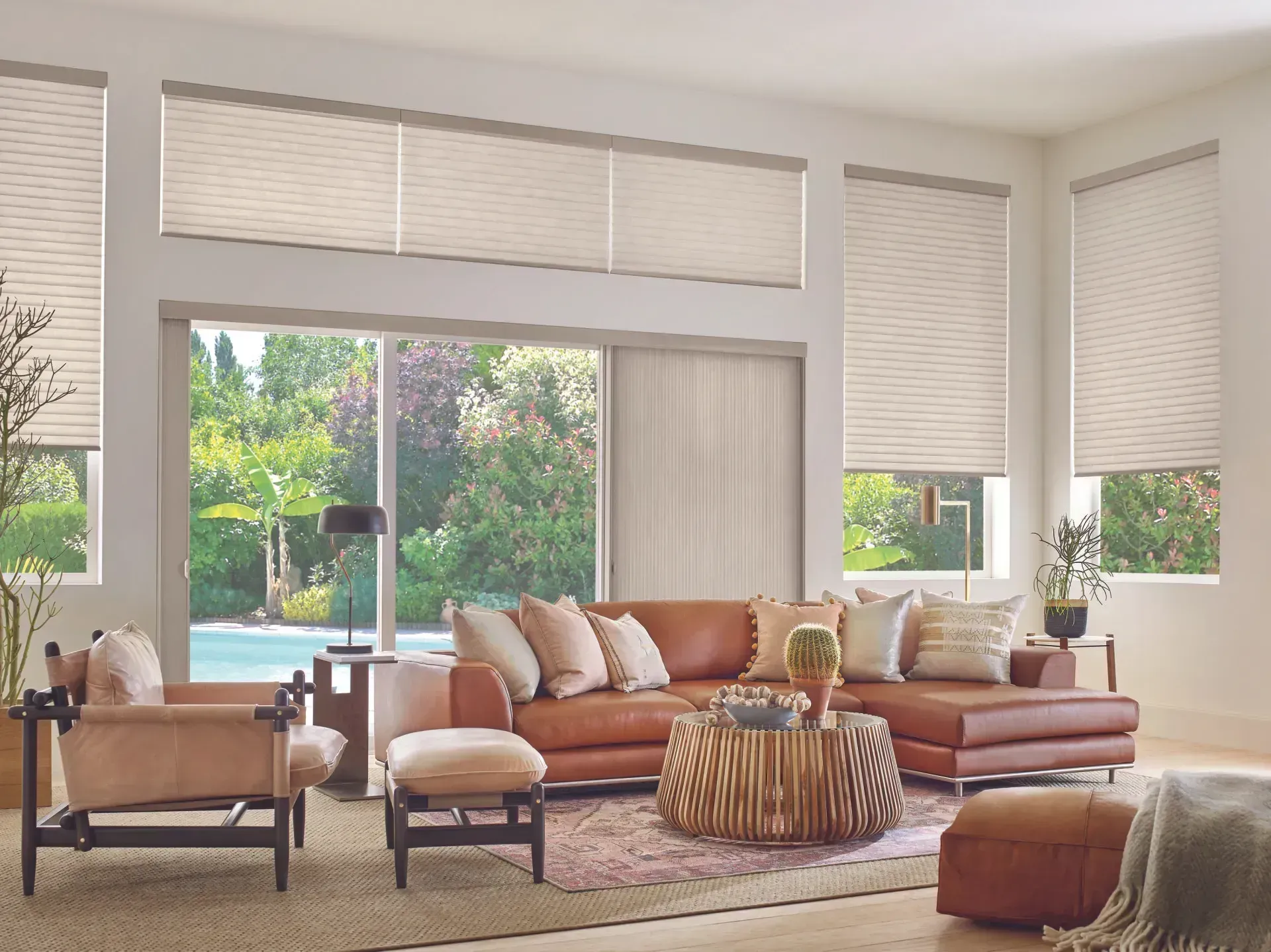 Living room with tan blinds, a brown leather sofa, and a view of a lush backyard.