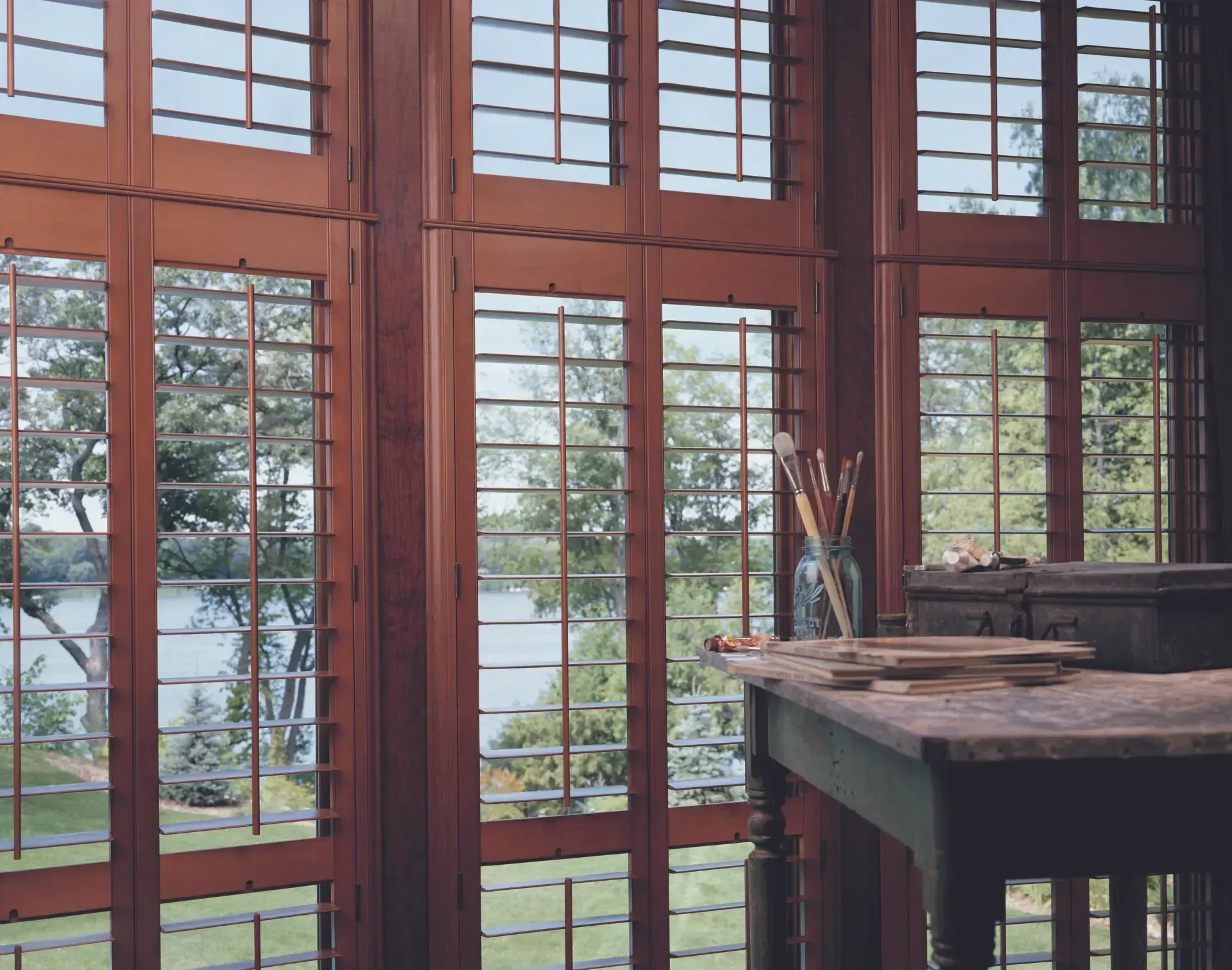 Wooden framed windows with grid patterns overlooking a lake. A desk with art supplies sits by the window.