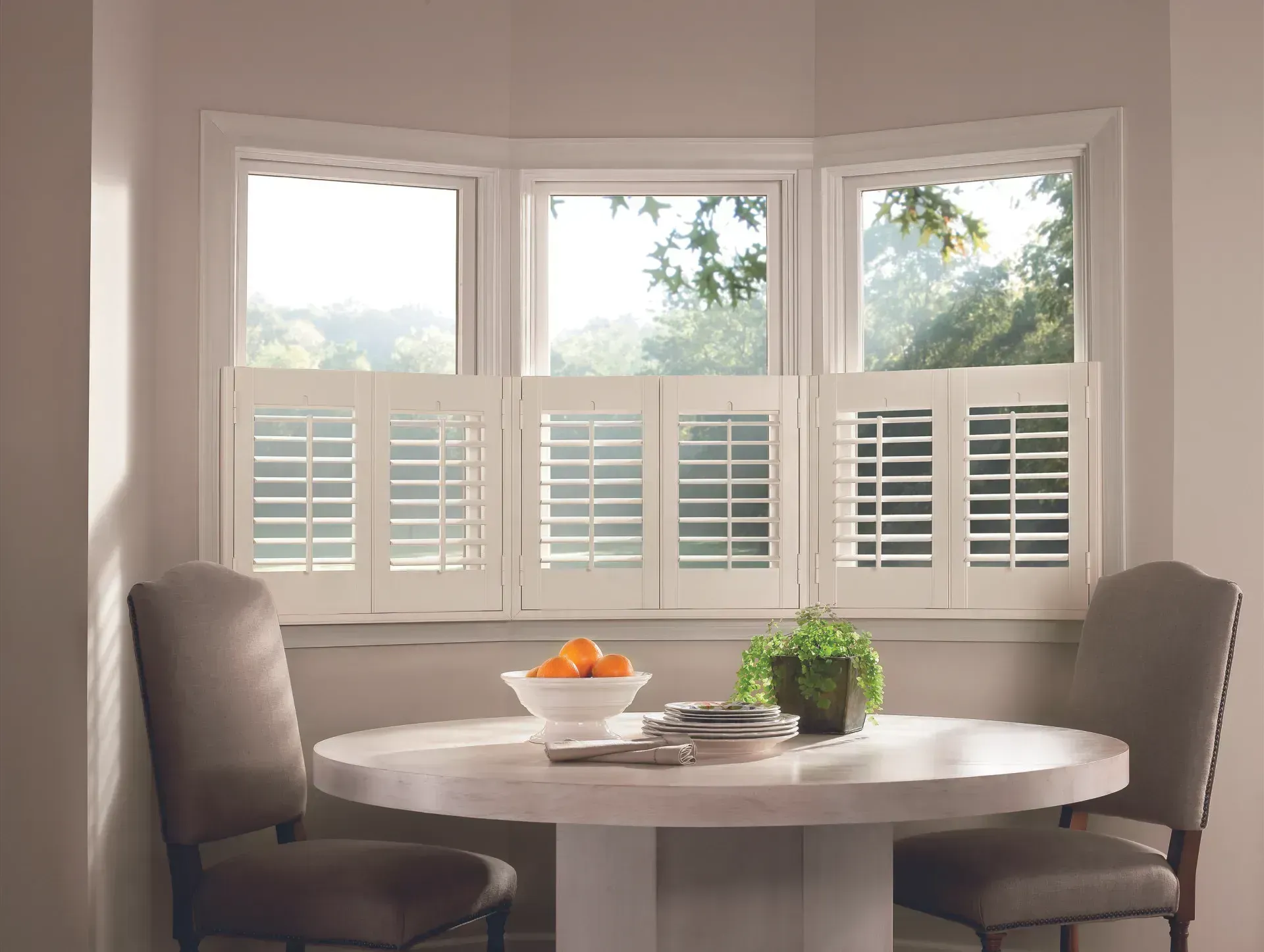 Dining room with round table, chairs, and bay window with white shutters.