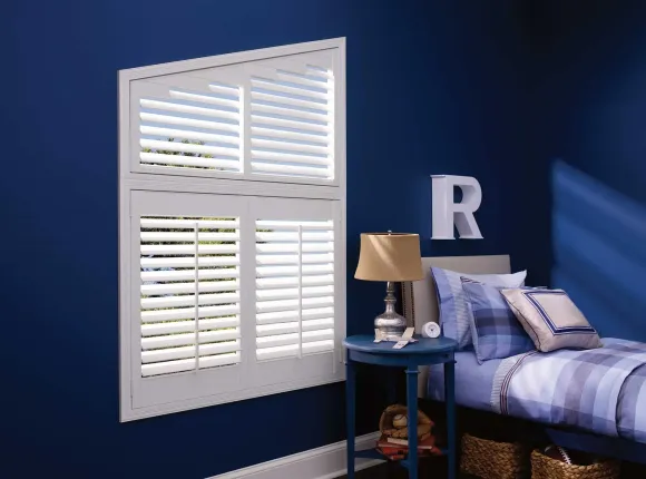 White shutters on a window in a blue-walled bedroom, beside a bed and table with lamp.