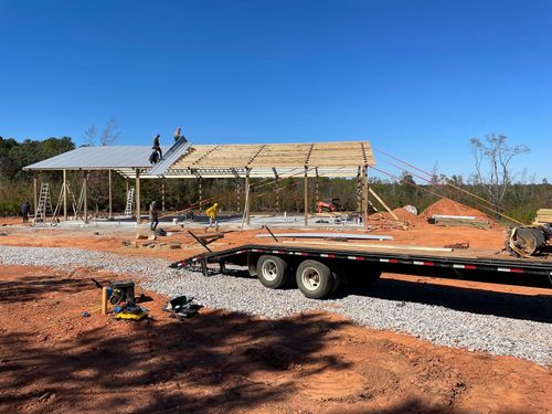 Construction site with workers on a metal roof frame beside a trailer and gravel driveway under a blue sky