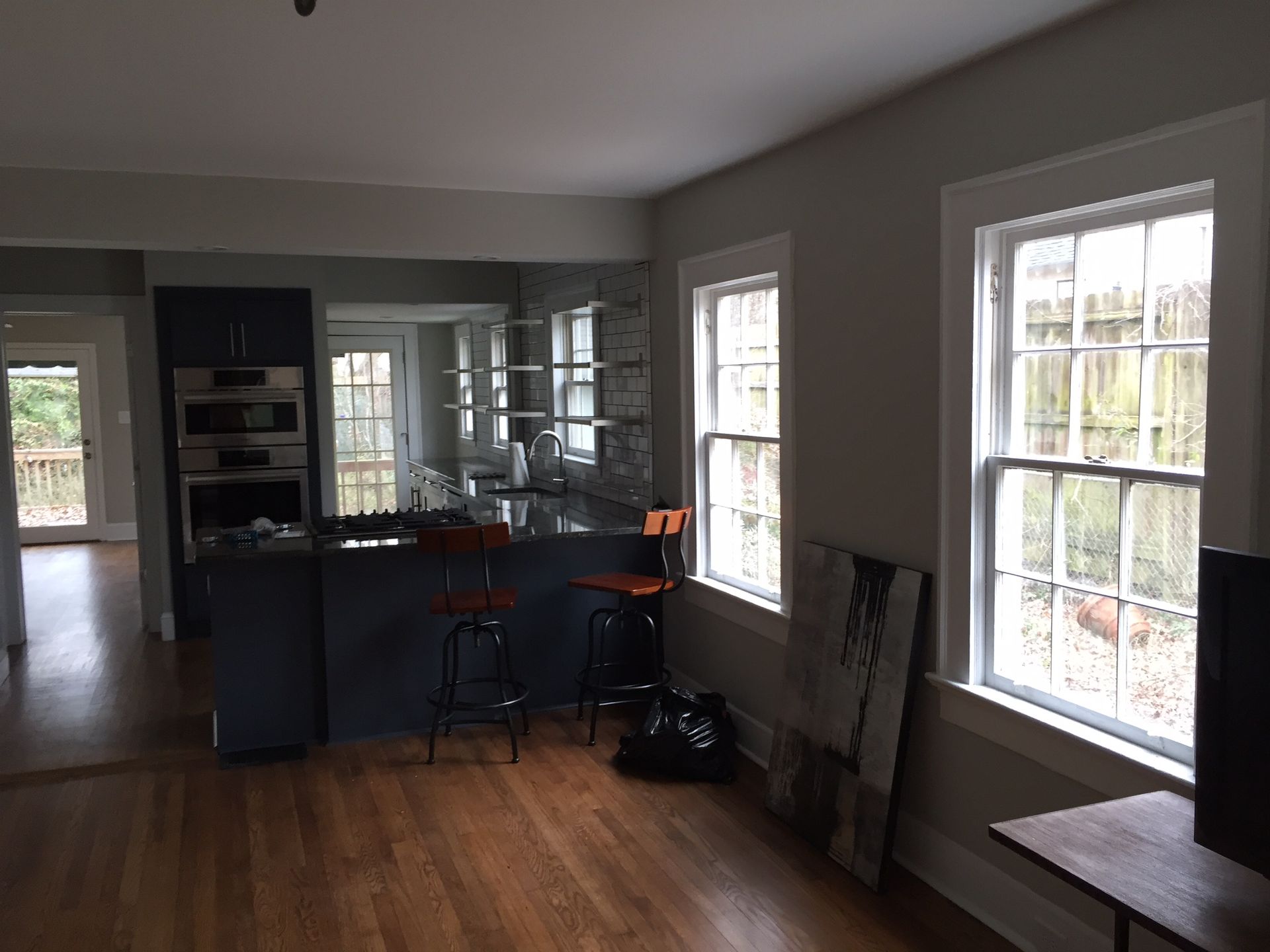 Empty living room with wood floors, gray walls, windows, and a dark kitchen island with stools.