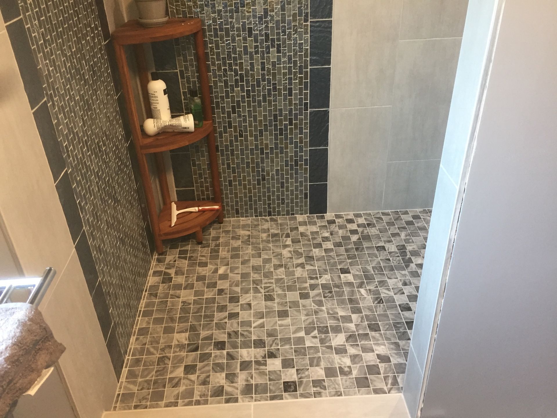 Small tiled shower with mosaic floor and wood corner shelf holding toiletries