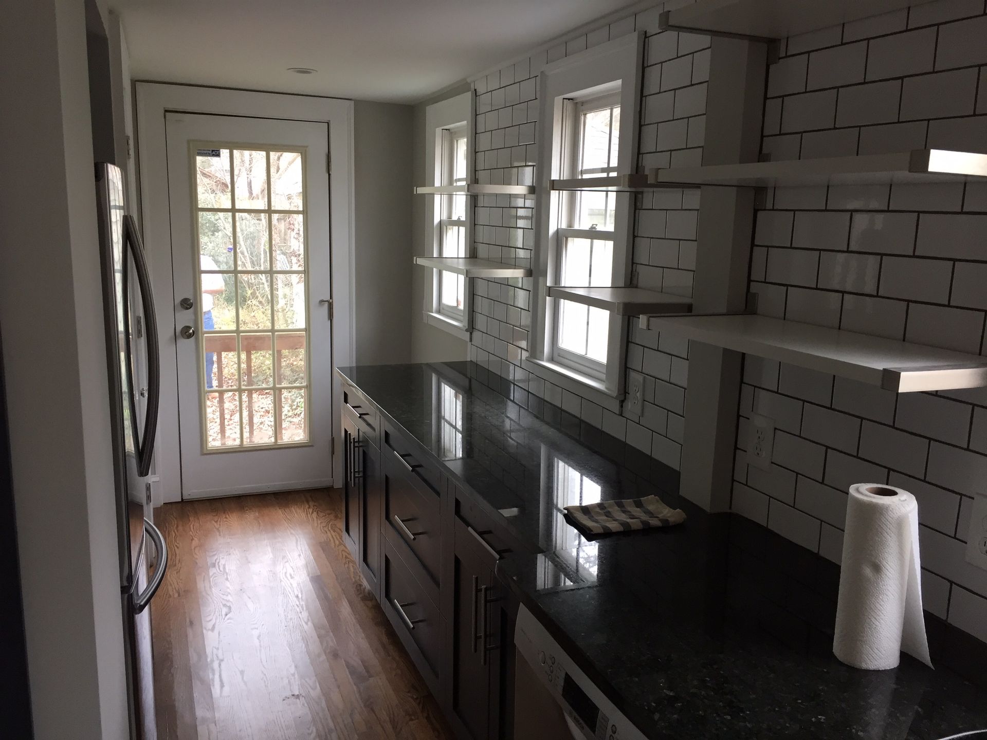Narrow kitchen with black countertops, white subway tile, wood floors, and a glass door at the end.