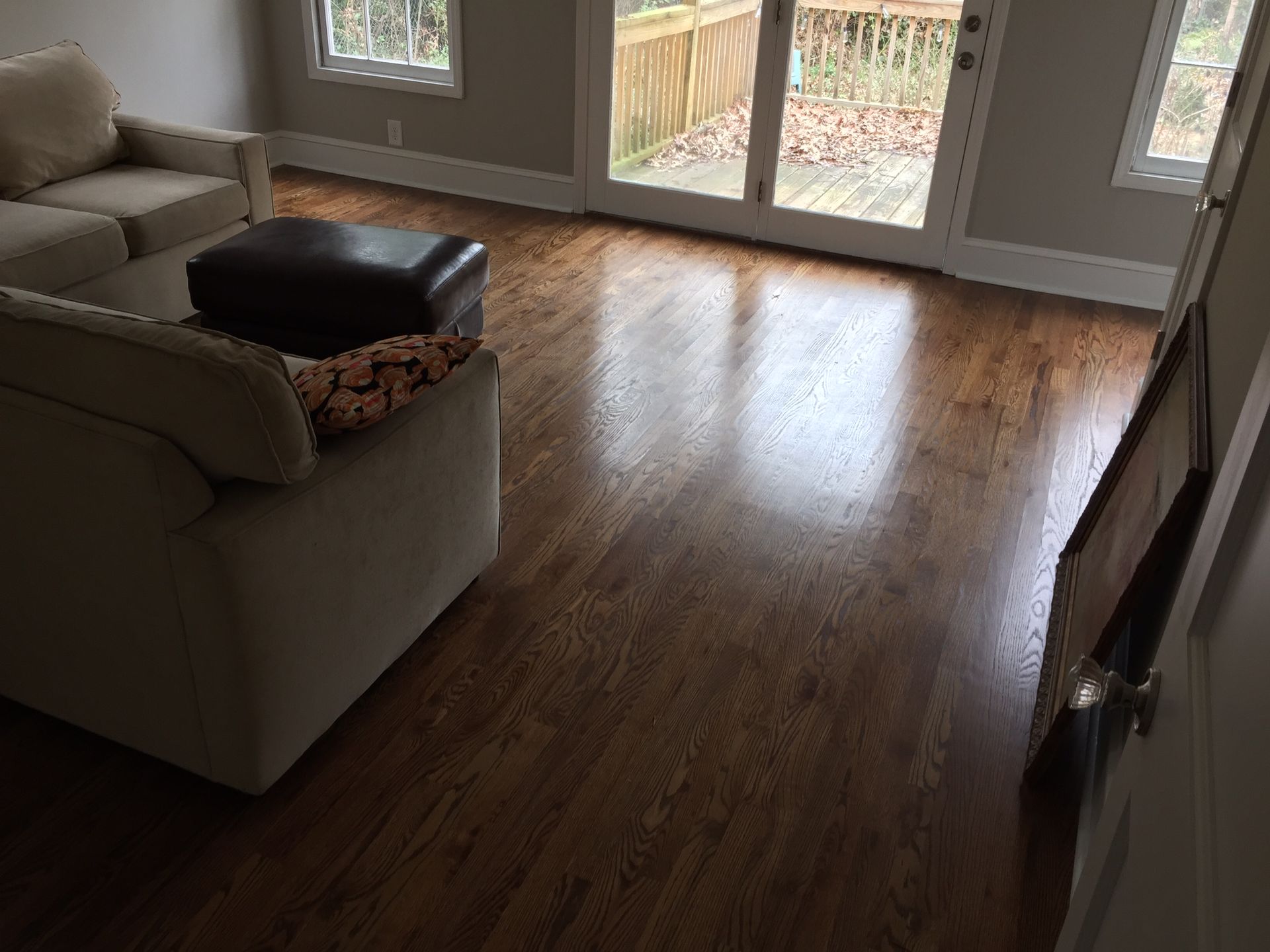 Empty living room with a beige couch, dark ottoman, and sunlight on a hardwood floor near glass doors.