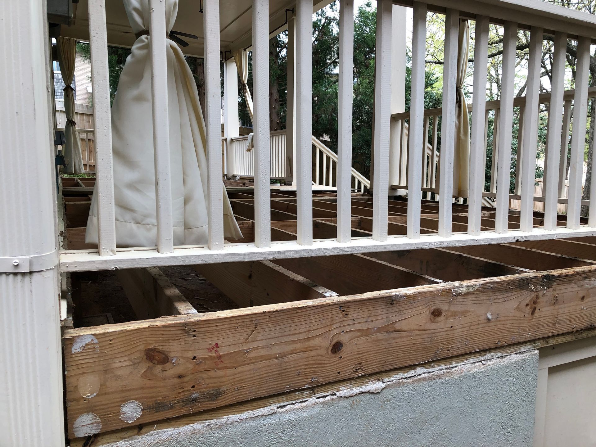 Damaged wooden porch with exposed joists and white railing, next to a house exterior