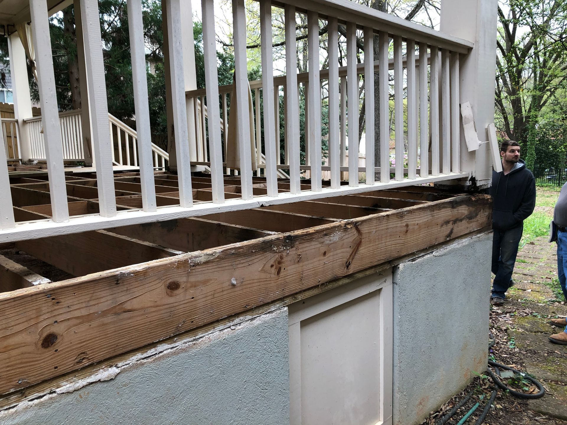 Damaged wooden porch deck with exposed joists beside a house, with a man standing nearby.