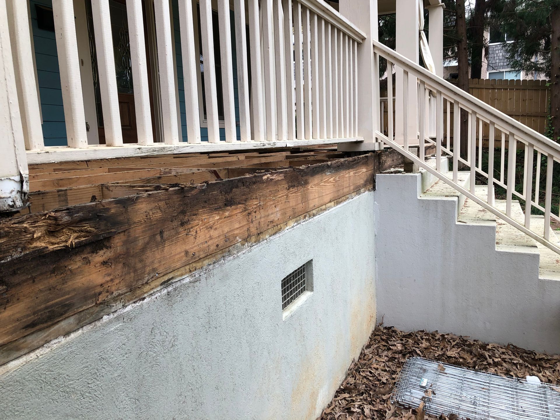 Damaged wooden porch railing and rot above a basement wall with a small vent and steps covered in leaves
