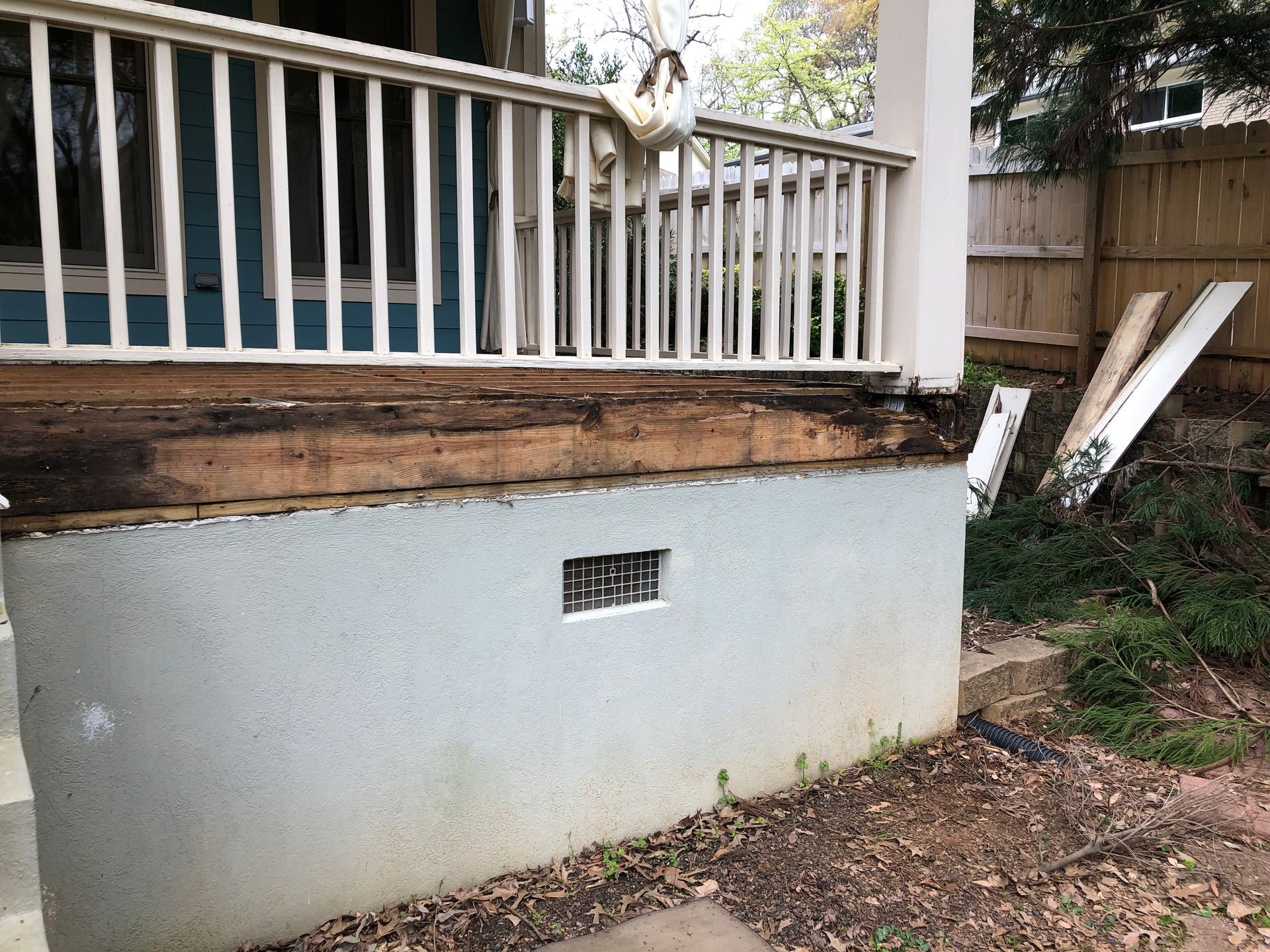 Raised porch with white railing and weathered foundation beside a yard with debris and wood planks
