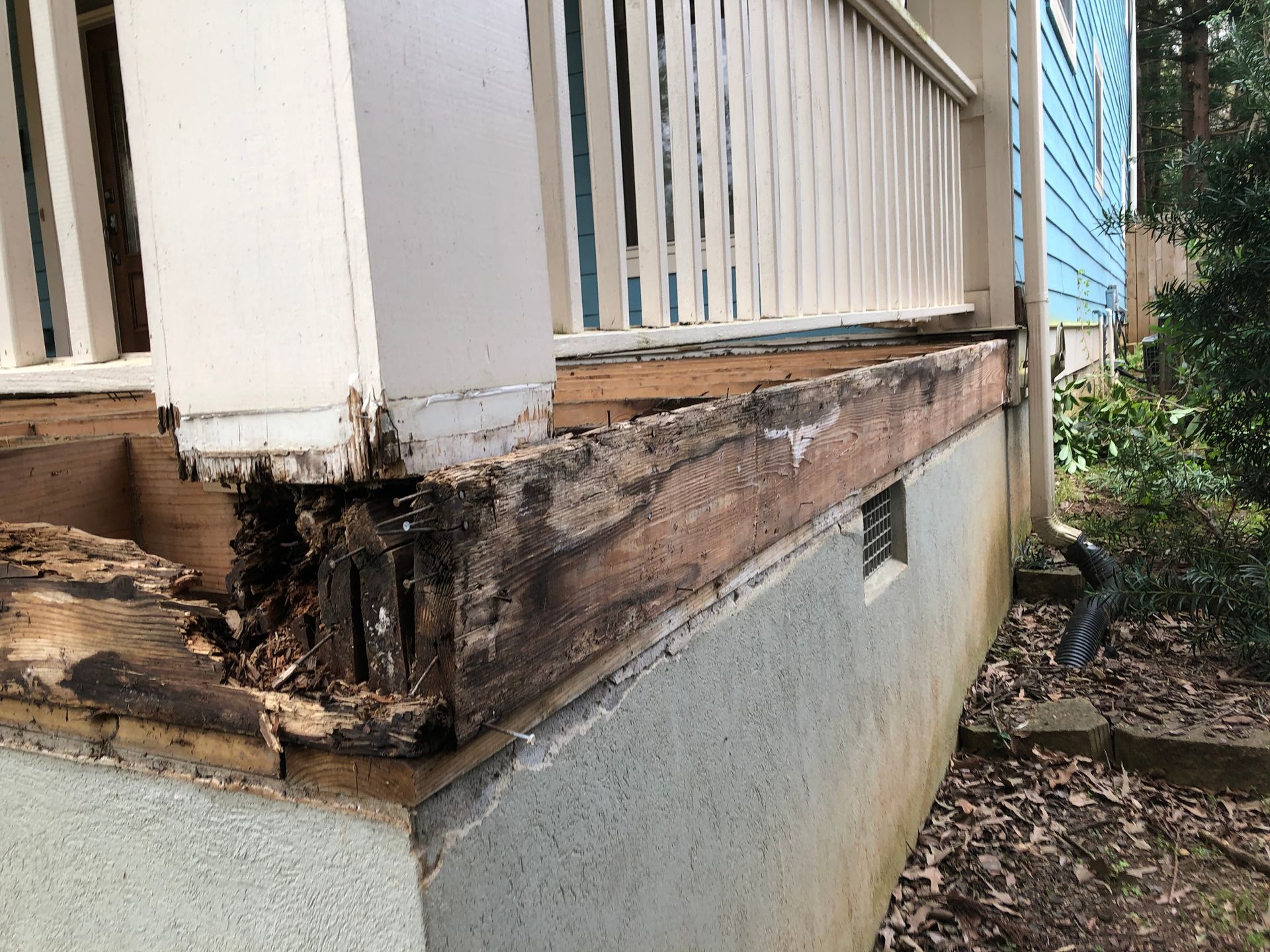 Rotting wooden porch support and siding on a house exterior, with visible water damage and decay