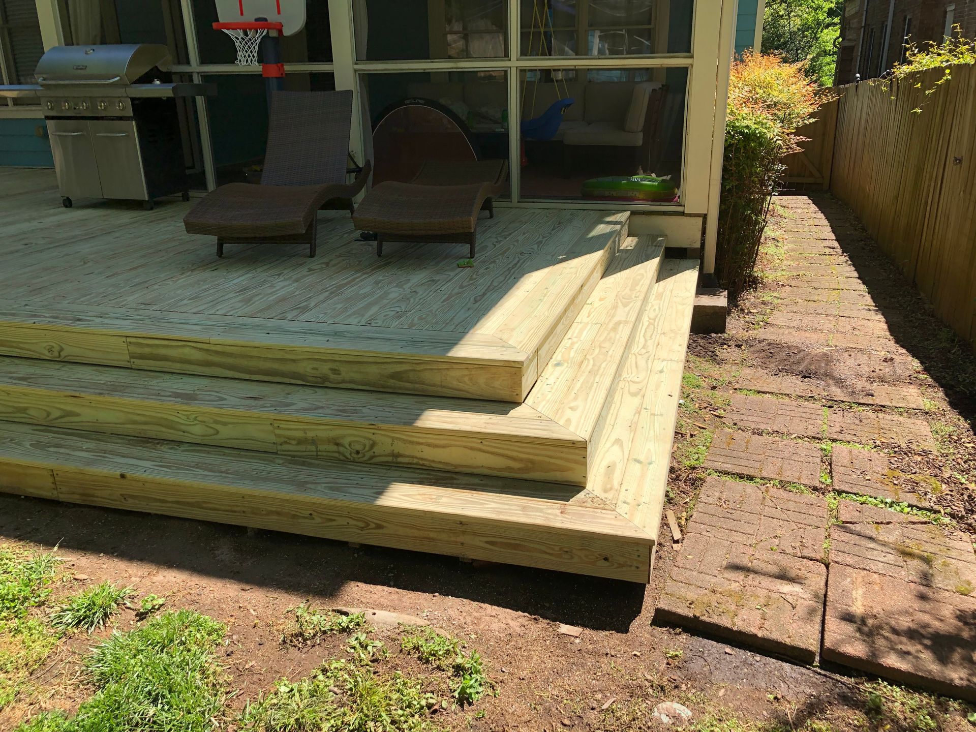 Raised wooden deck steps beside a patio with lounge chairs and a grill in a backyard.